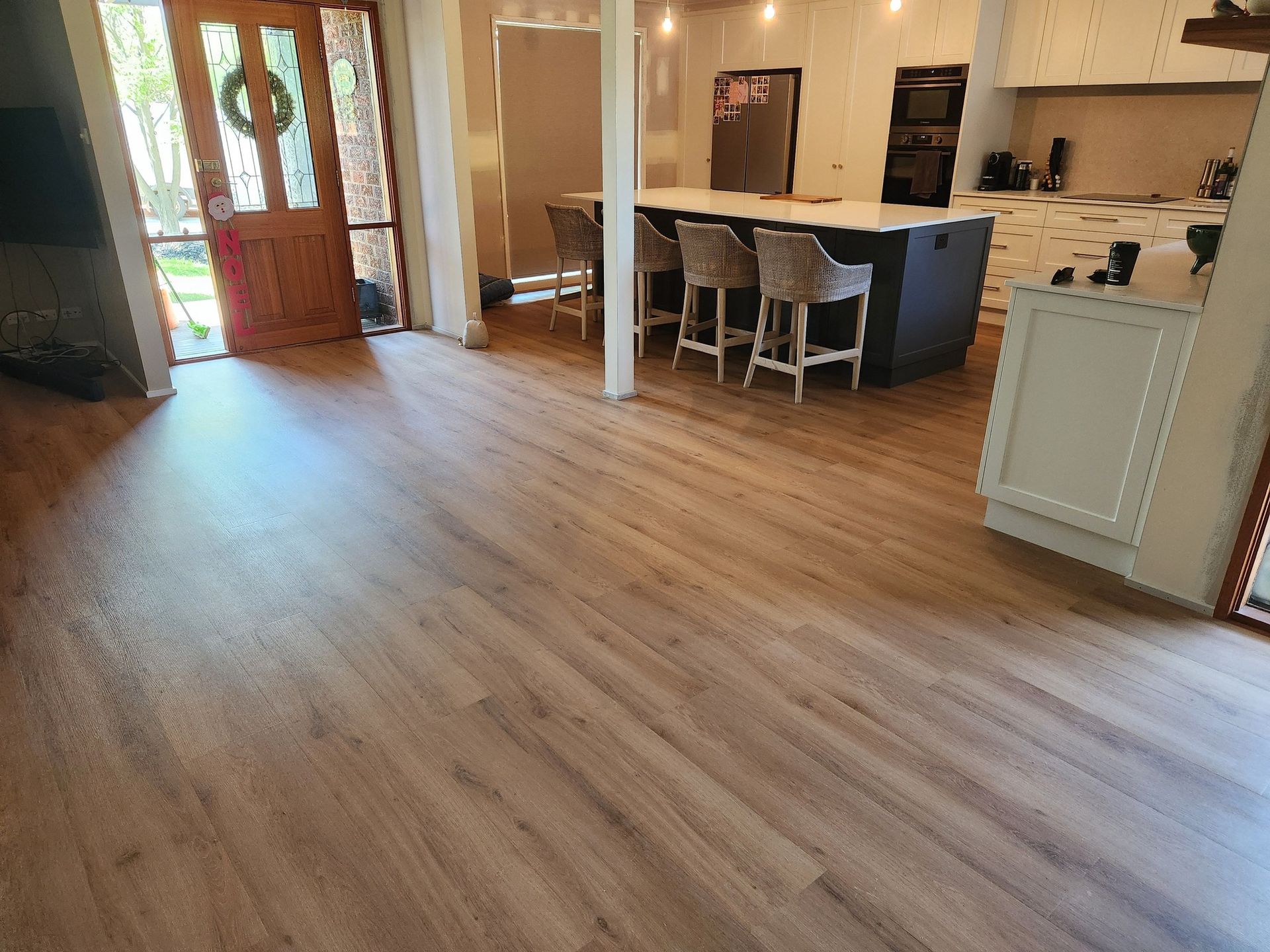 Open-Concept Kitchen and Living Area with Wood-Look Flooring, Kitchen Island with Barstools, and A Front Door — Canberra Floor Coverings in Queanbeyan West, NSW