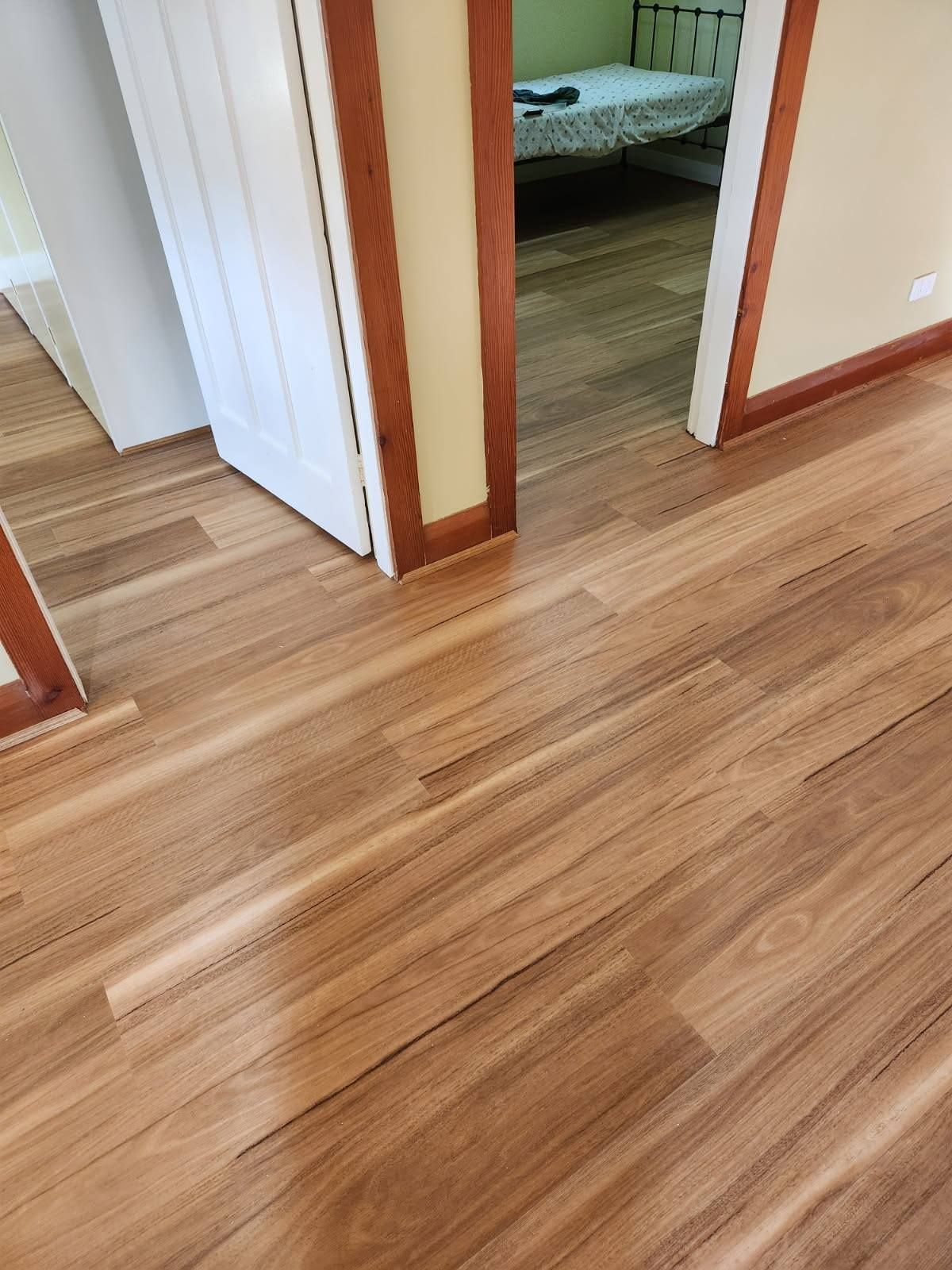 Wooden Floor Leading to A Doorway with Light Brown Trim and A Room with A Bed — Canberra Floor Coverings in Queanbeyan West, NSW