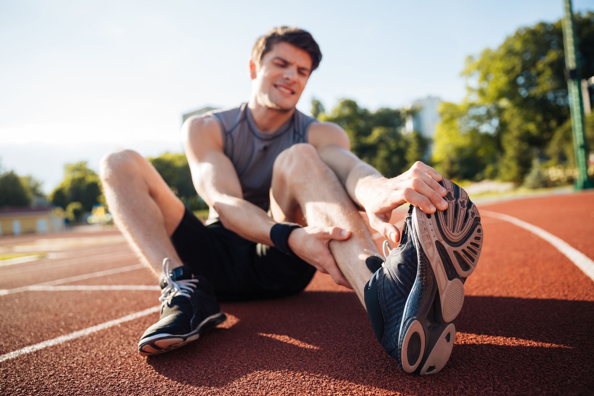 Man seated on track, holding ankle in pain; wearing athletic gear.