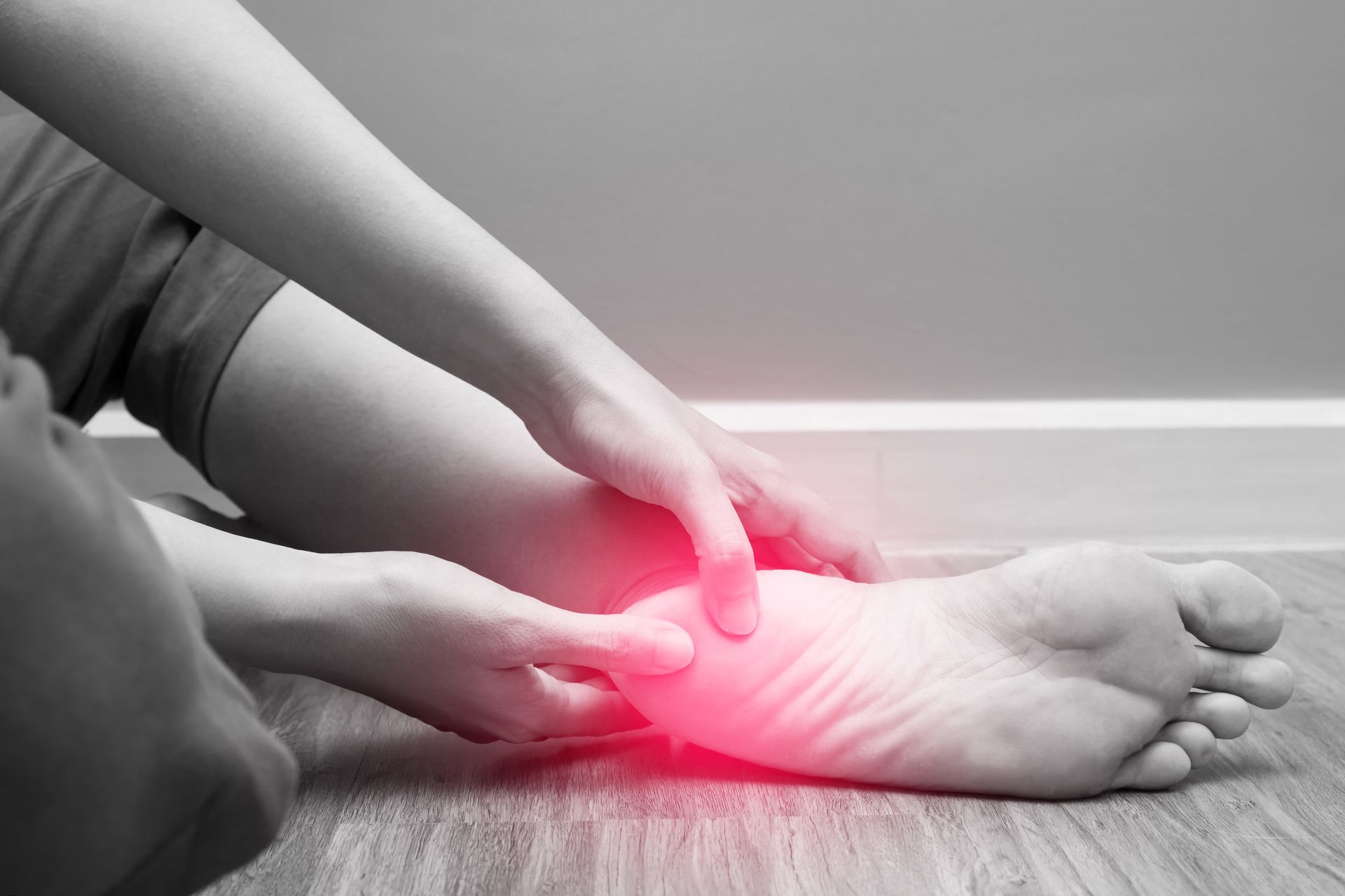 Person holding their foot, highlighting heel pain with a red glow, seated on wooden floor.