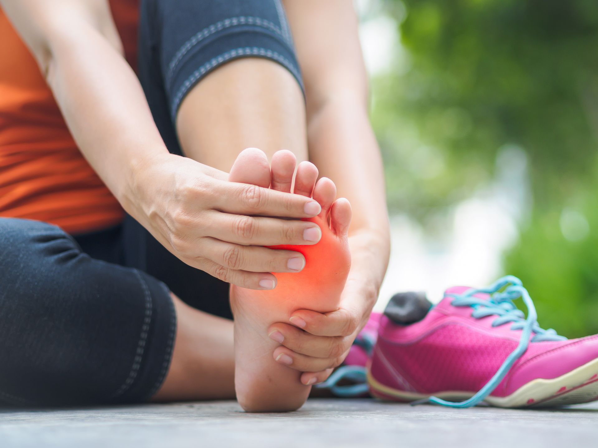 Woman seated, holding foot with red highlighted pain area. Pink shoe next to her. Outdoor setting.