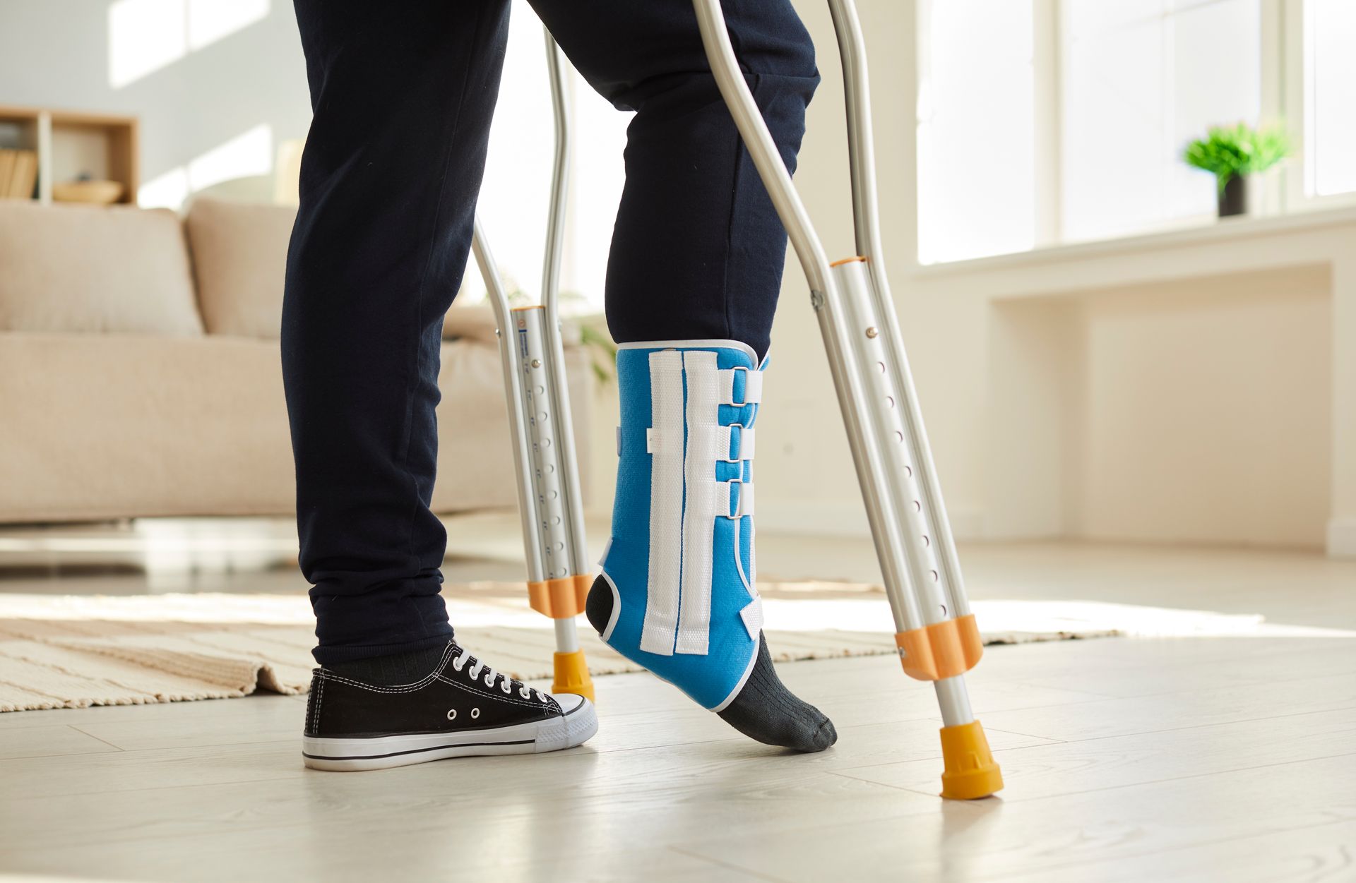 Person using crutches, leg in a blue medical boot. Indoors, walking on a hardwood floor.
