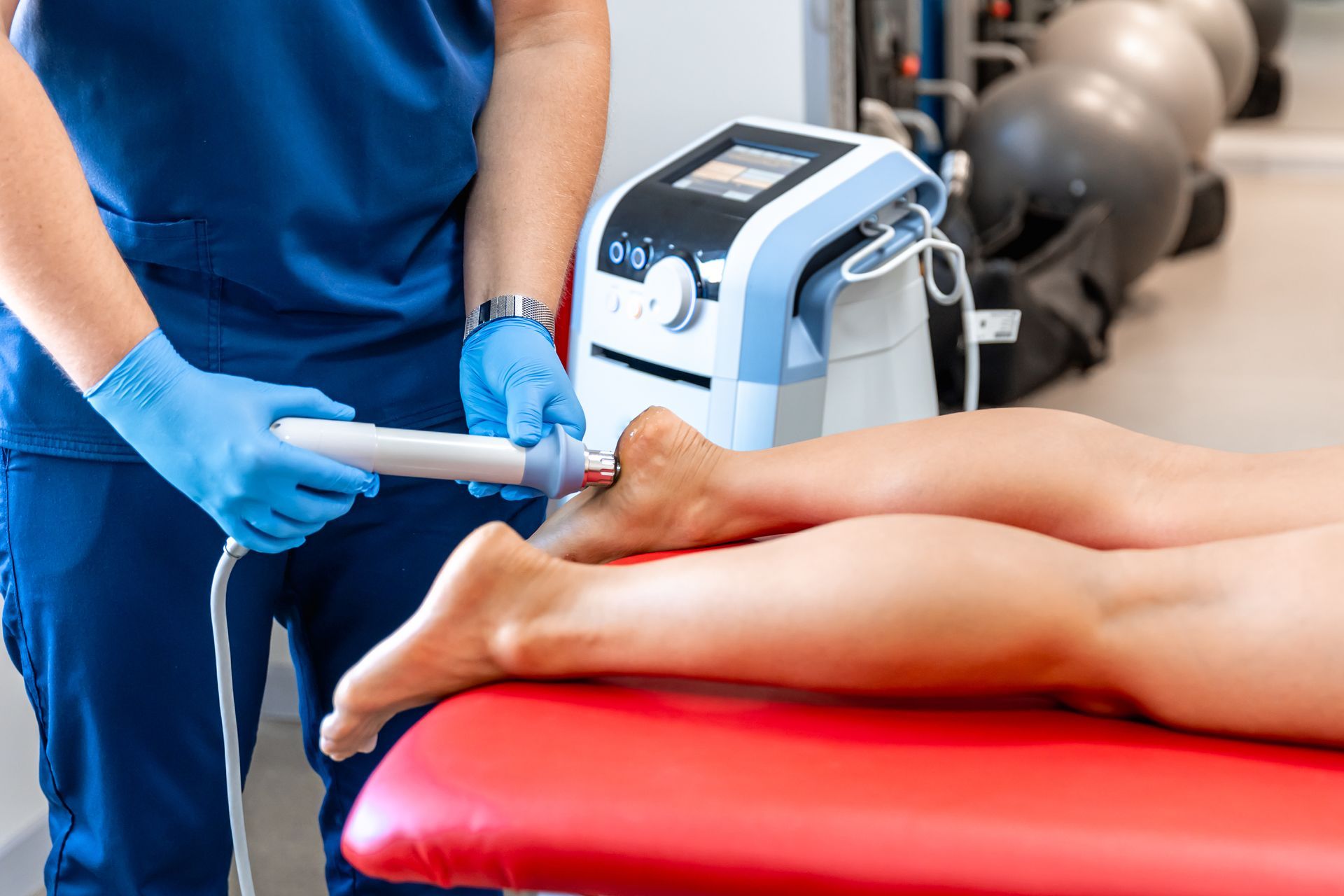 Physician treating a patient's foot with a medical device. Blue scrubs, gloves, white device, red table.