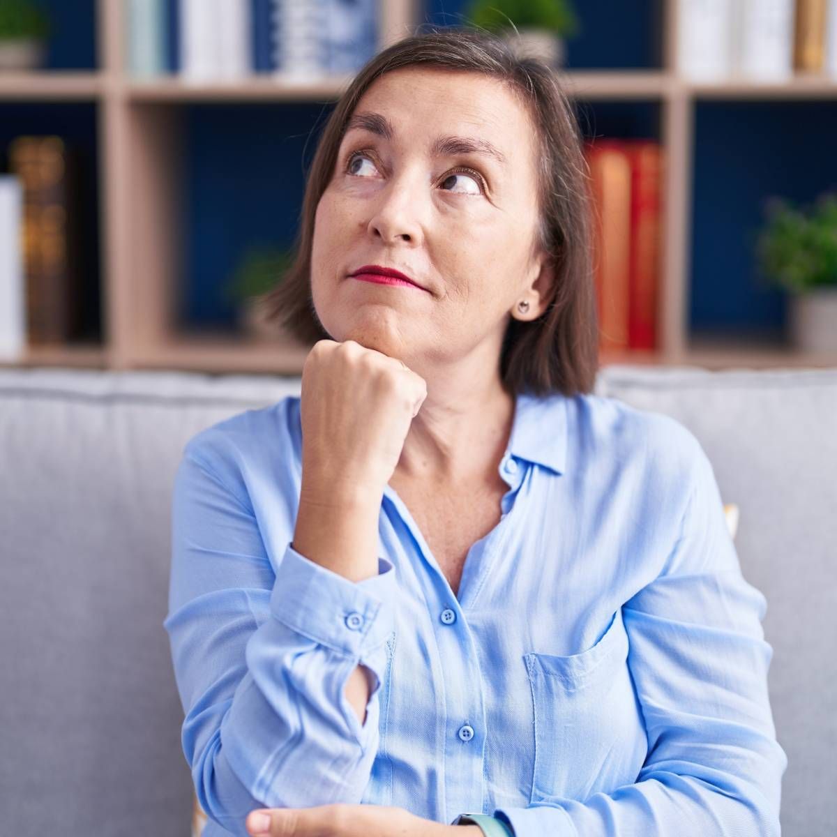 Woman in a blue shirt, looking upward, resting chin on hand, pondering. Background is a bookshelf.