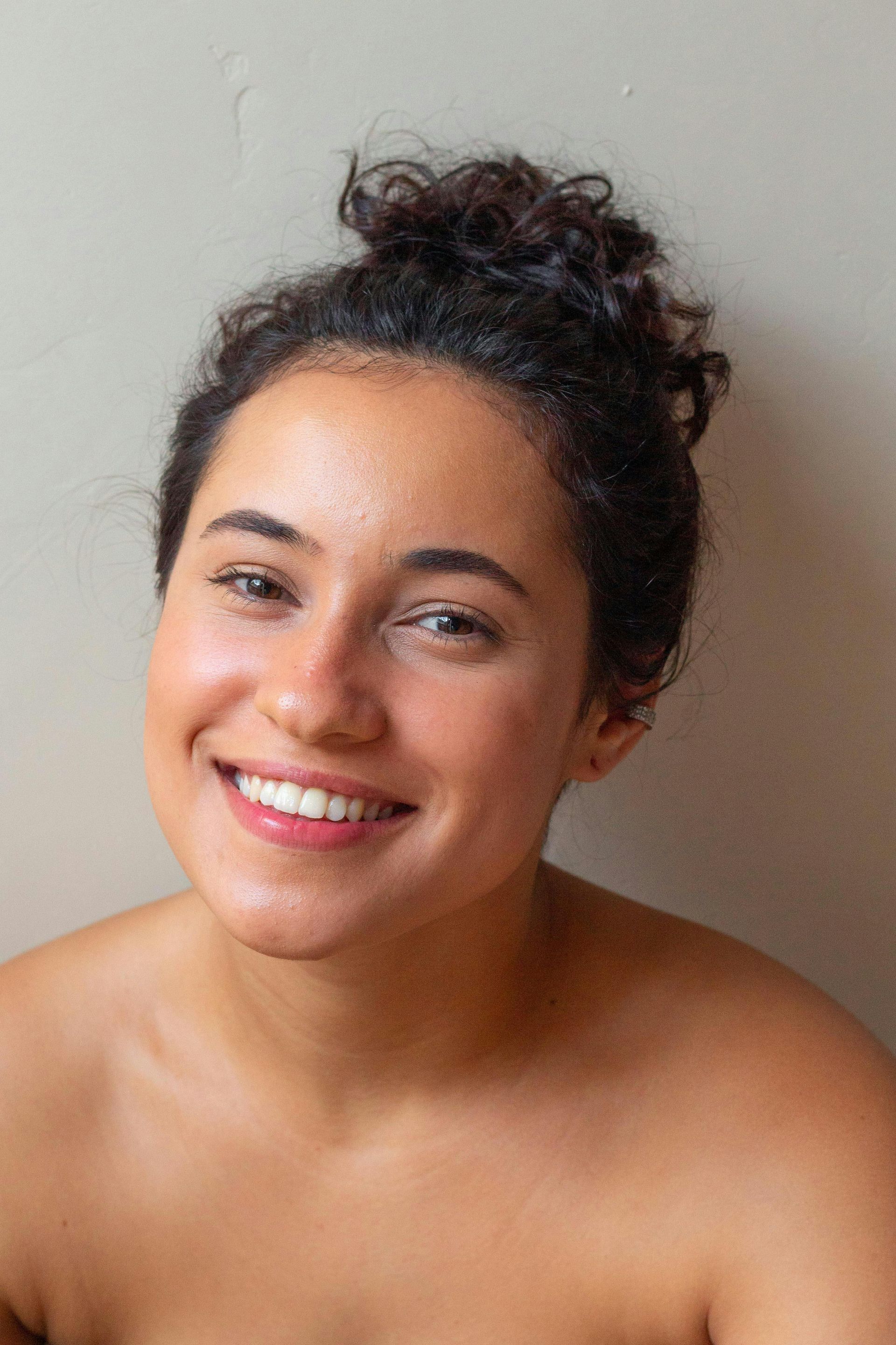 Woman with dark curly hair in a bun smiling widely, bare shoulders, against a neutral-toned wall.