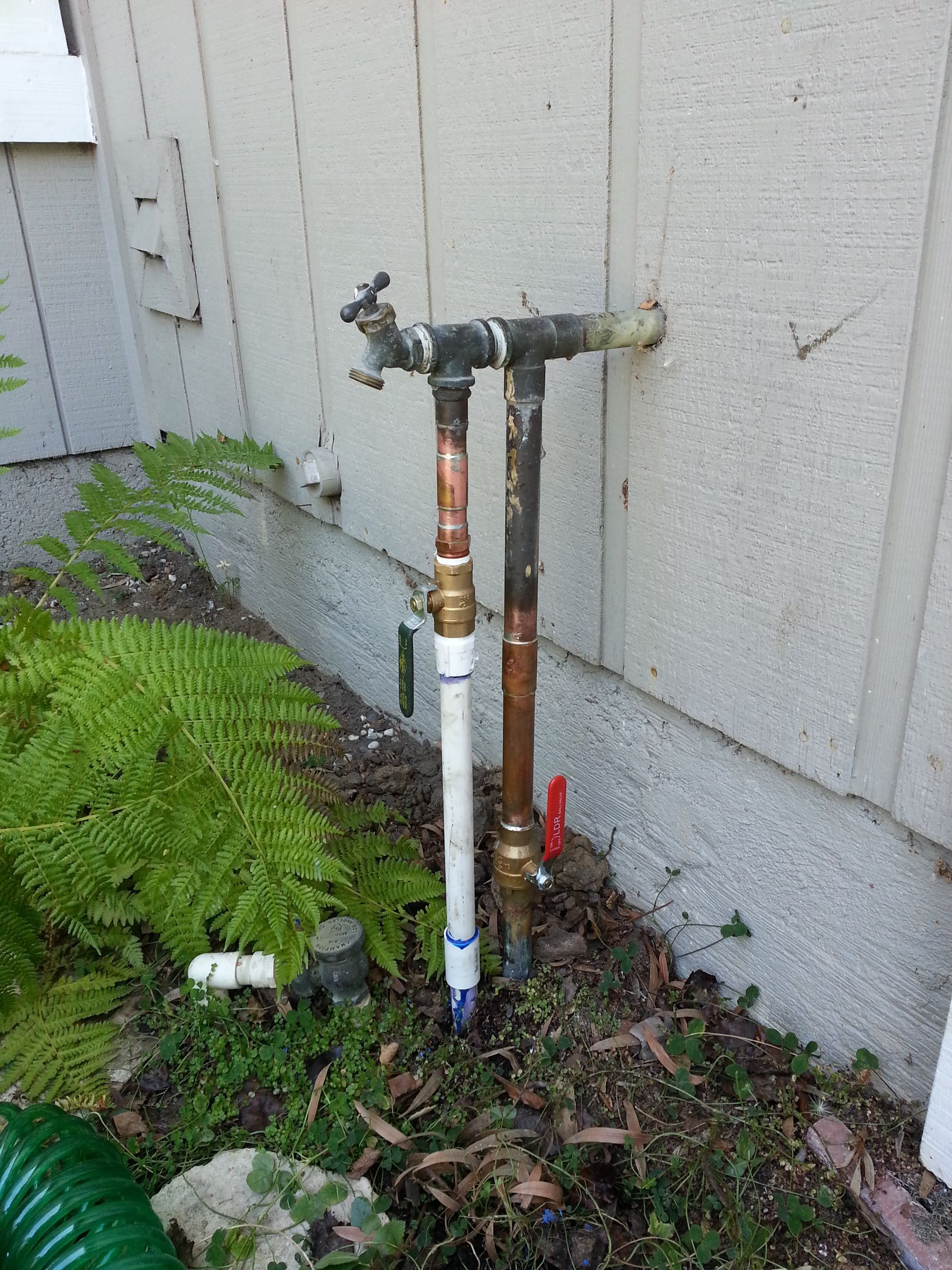 Outdoor water pipes and spigots attached to a beige house, next to greenery.