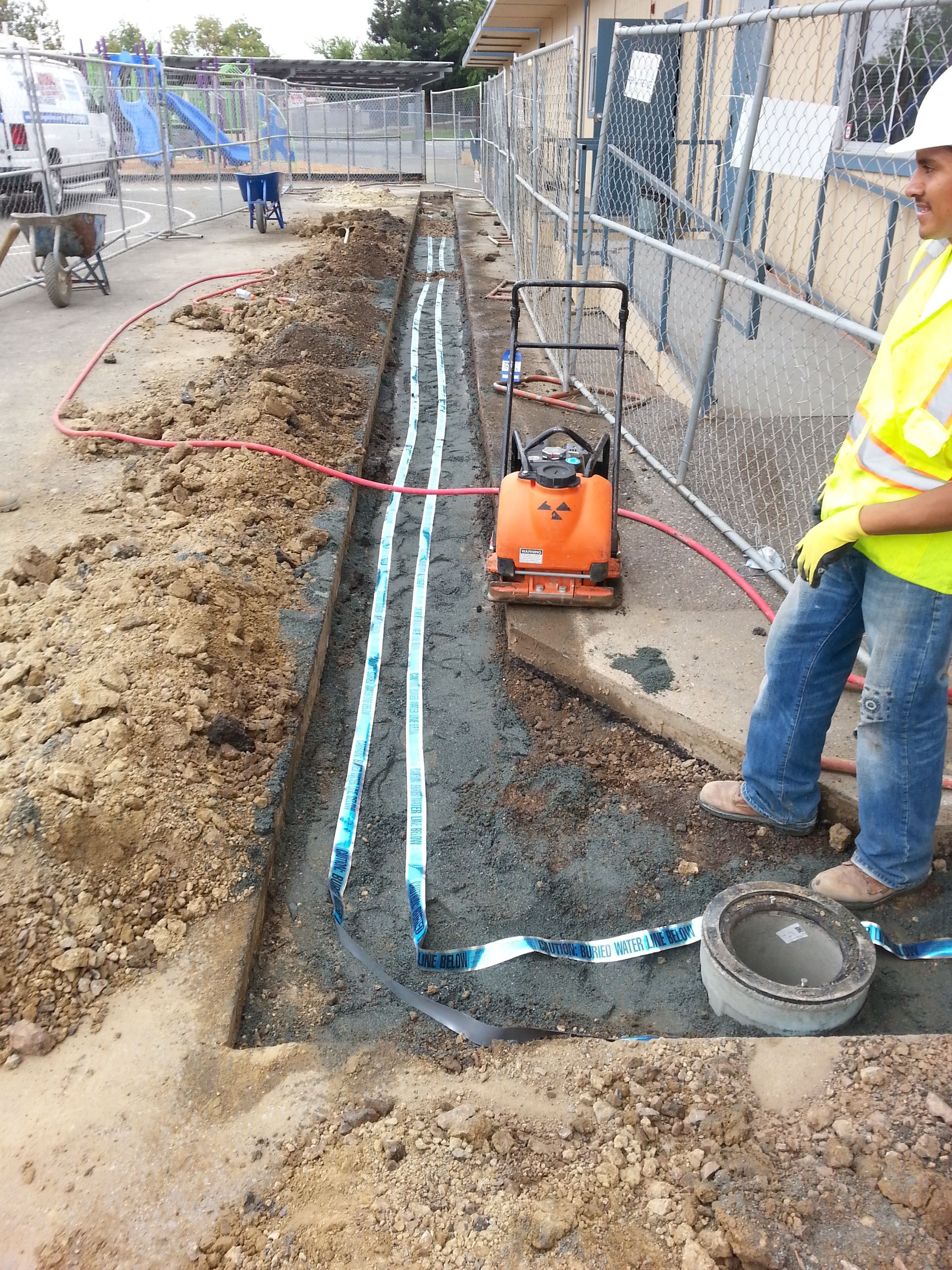 Construction worker beside trench with pipes, using compactor, near a building.