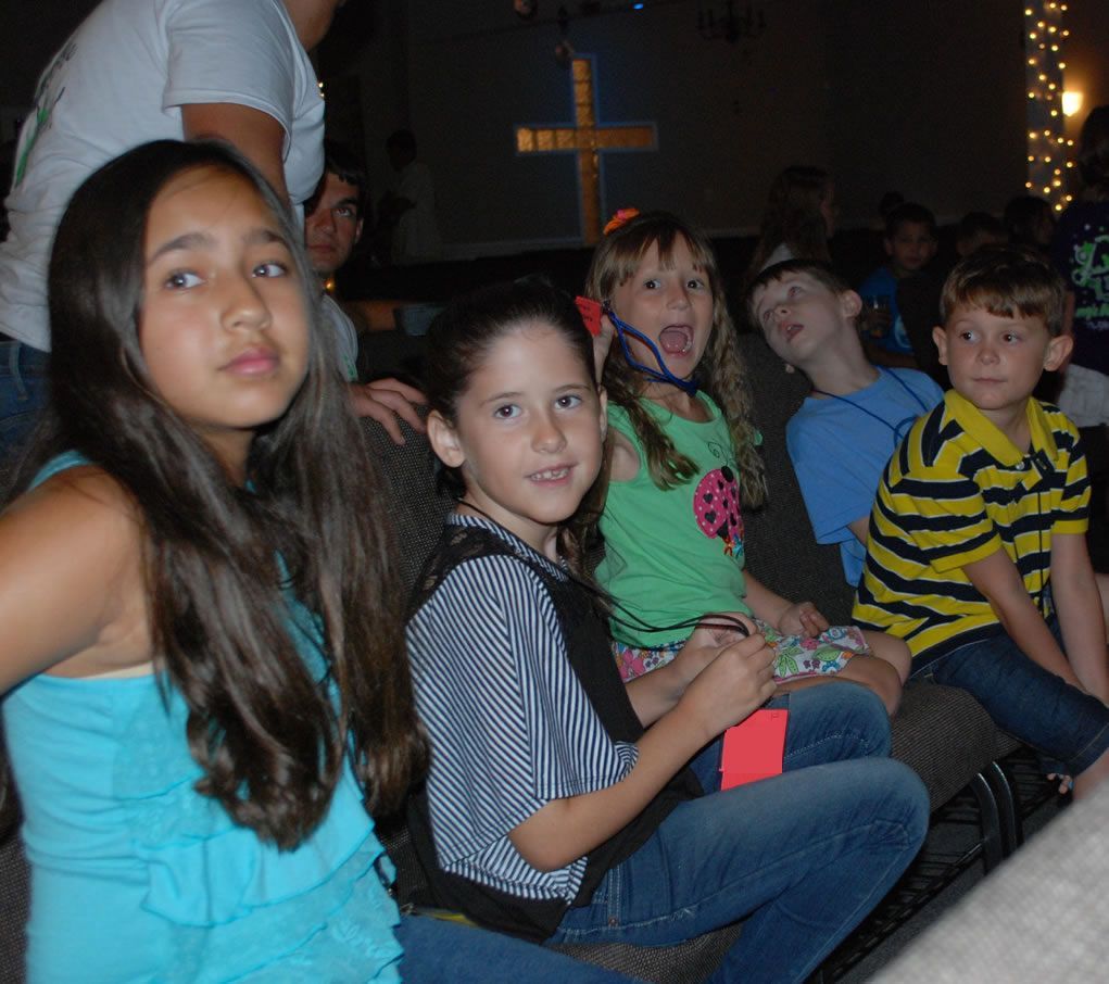 A group of children are sitting in a room with a cross in the background