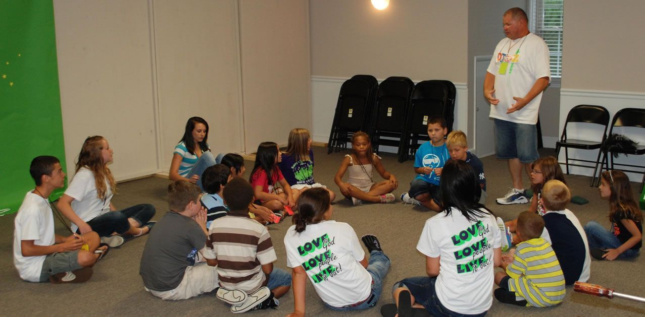 A group of children are sitting in a circle with a man standing in the middle.