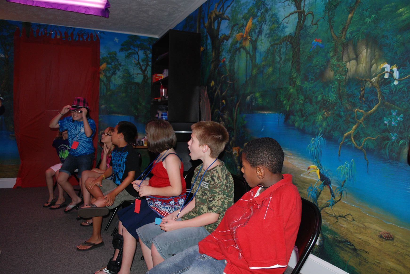 A group of children are sitting in a room with a painting on the wall.