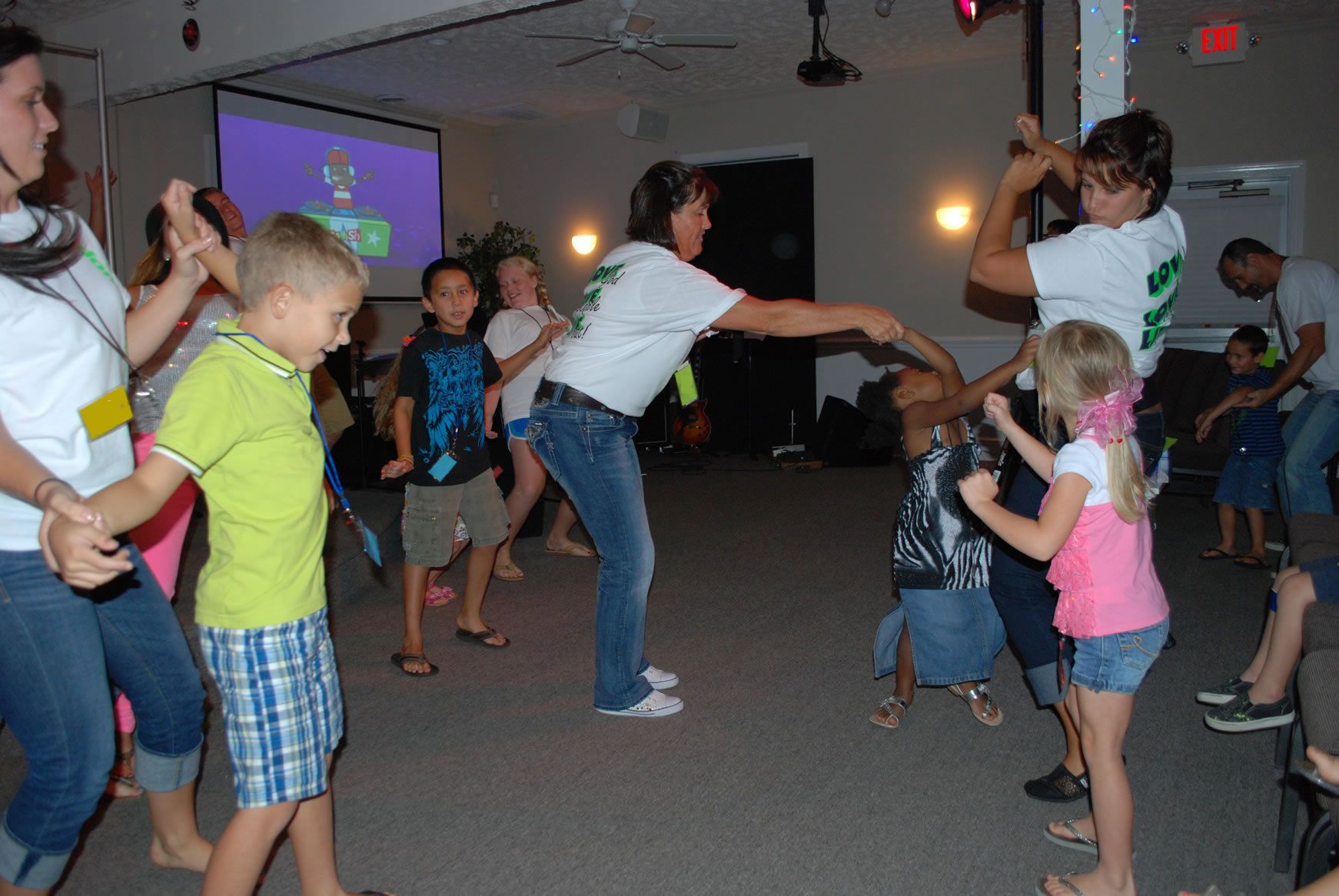 A group of people are dancing in a room with a projector screen in the background.