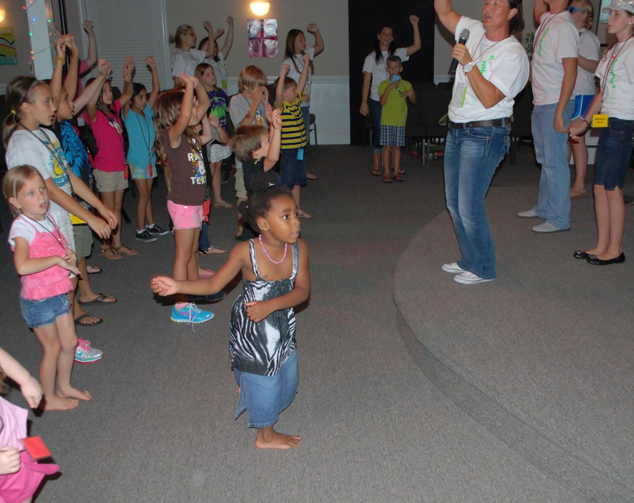 A group of children are dancing in a room with a man holding a microphone