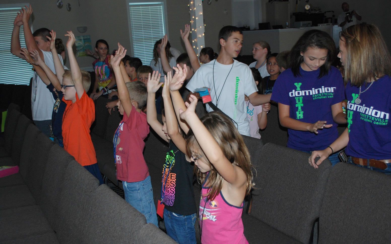 A group of children wearing dominion shirts are raising their hands in the air