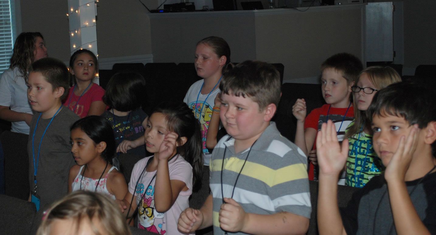 A group of children are sitting in a room raising their hands.