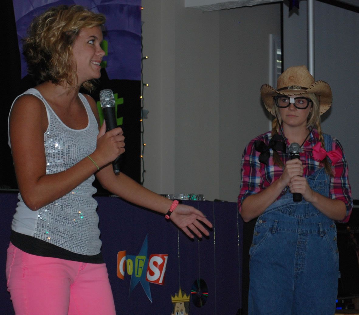 A girl in a cowboy hat stands next to a girl in overalls holding a microphone