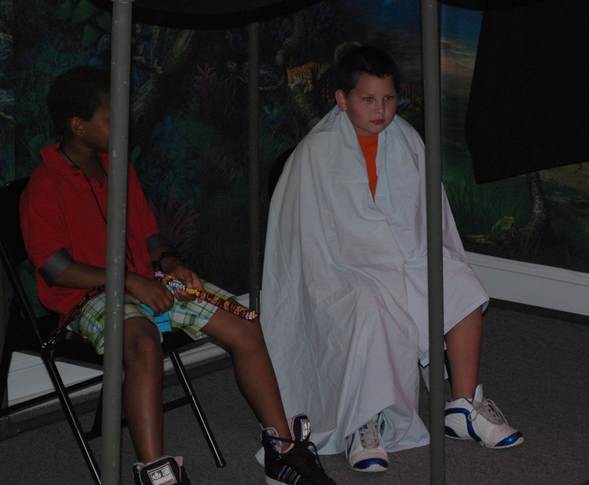 A boy wrapped in a white blanket sits next to another boy