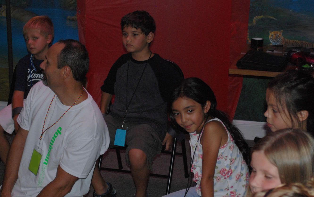 A group of children are sitting around a man wearing a lanyard that says leadership
