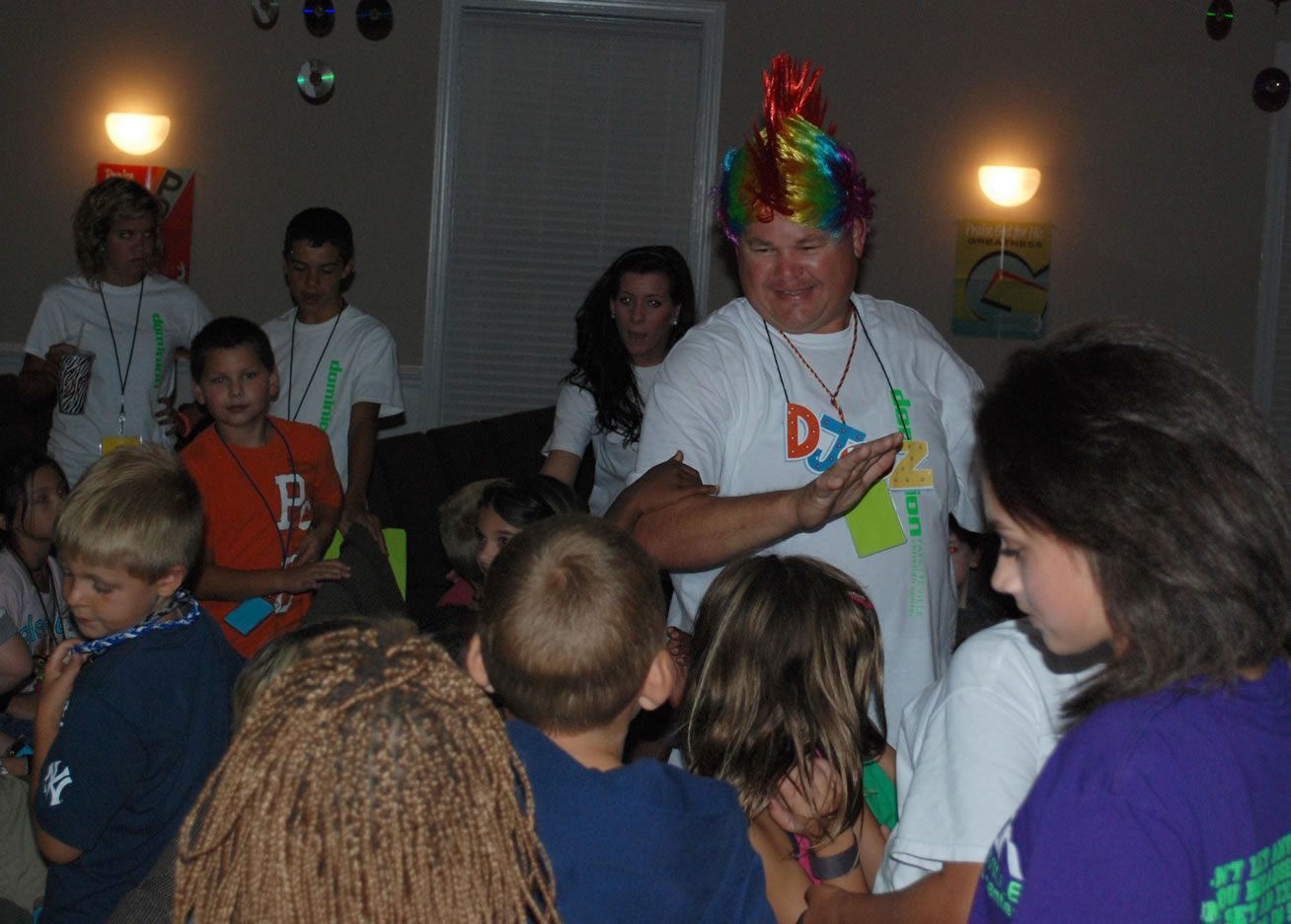 A man wearing a rainbow wig is standing in front of a group of children.