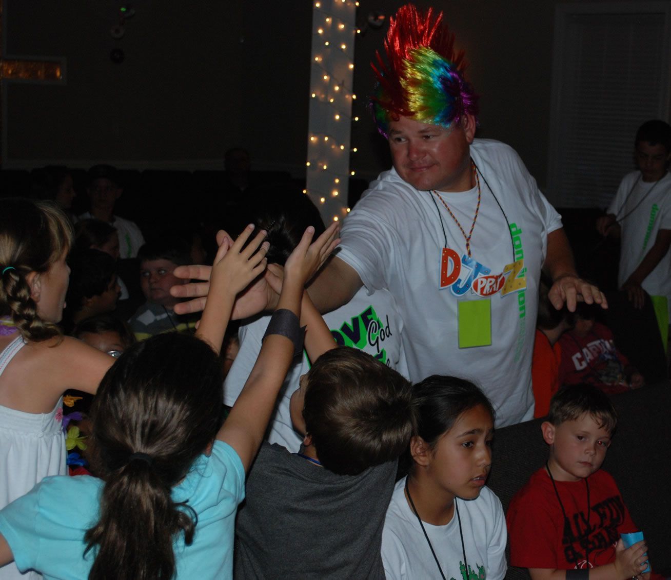 A man wearing a rainbow wig is surrounded by children