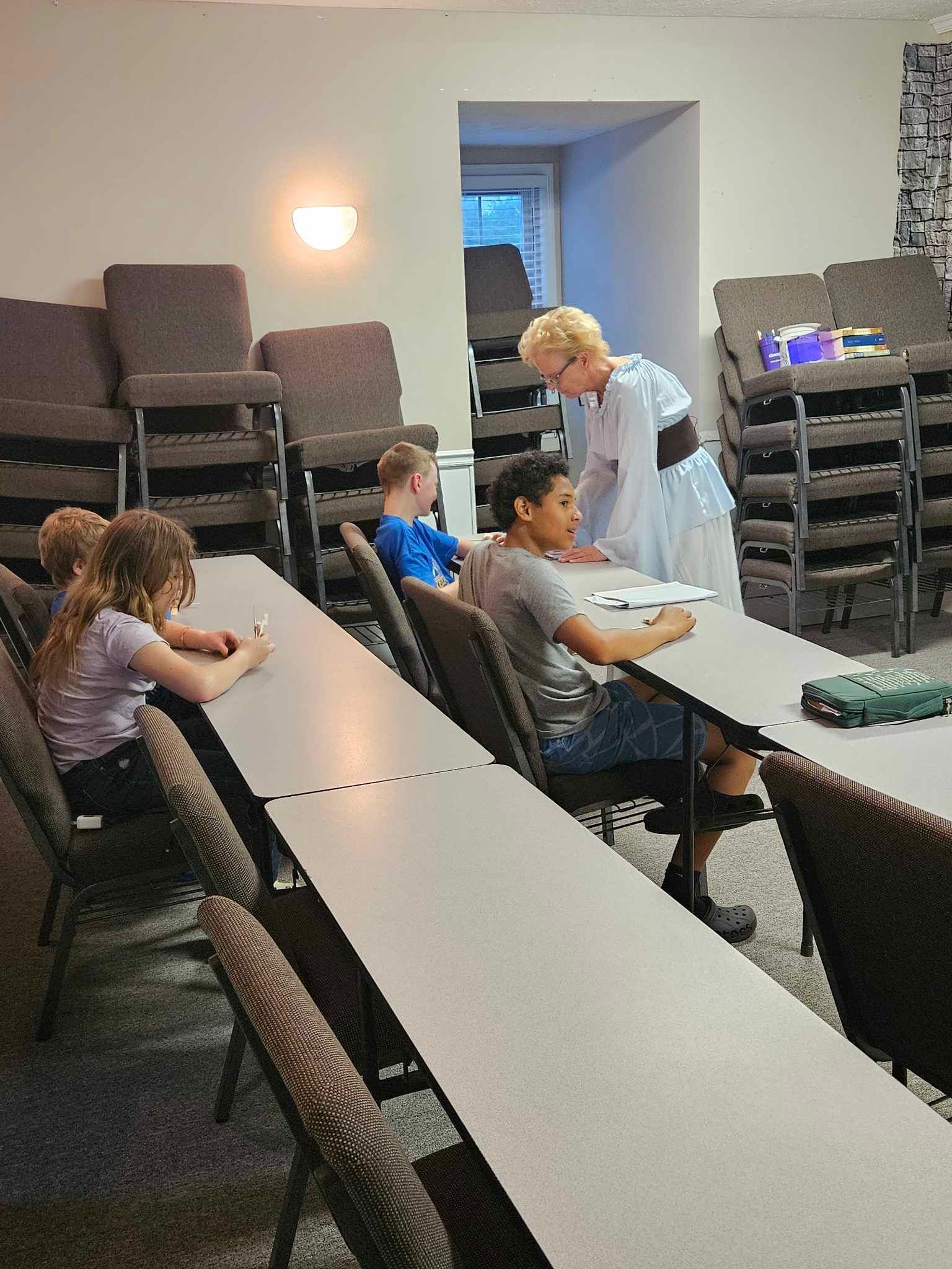 A woman is talking to a group of children sitting at tables in a classroom.