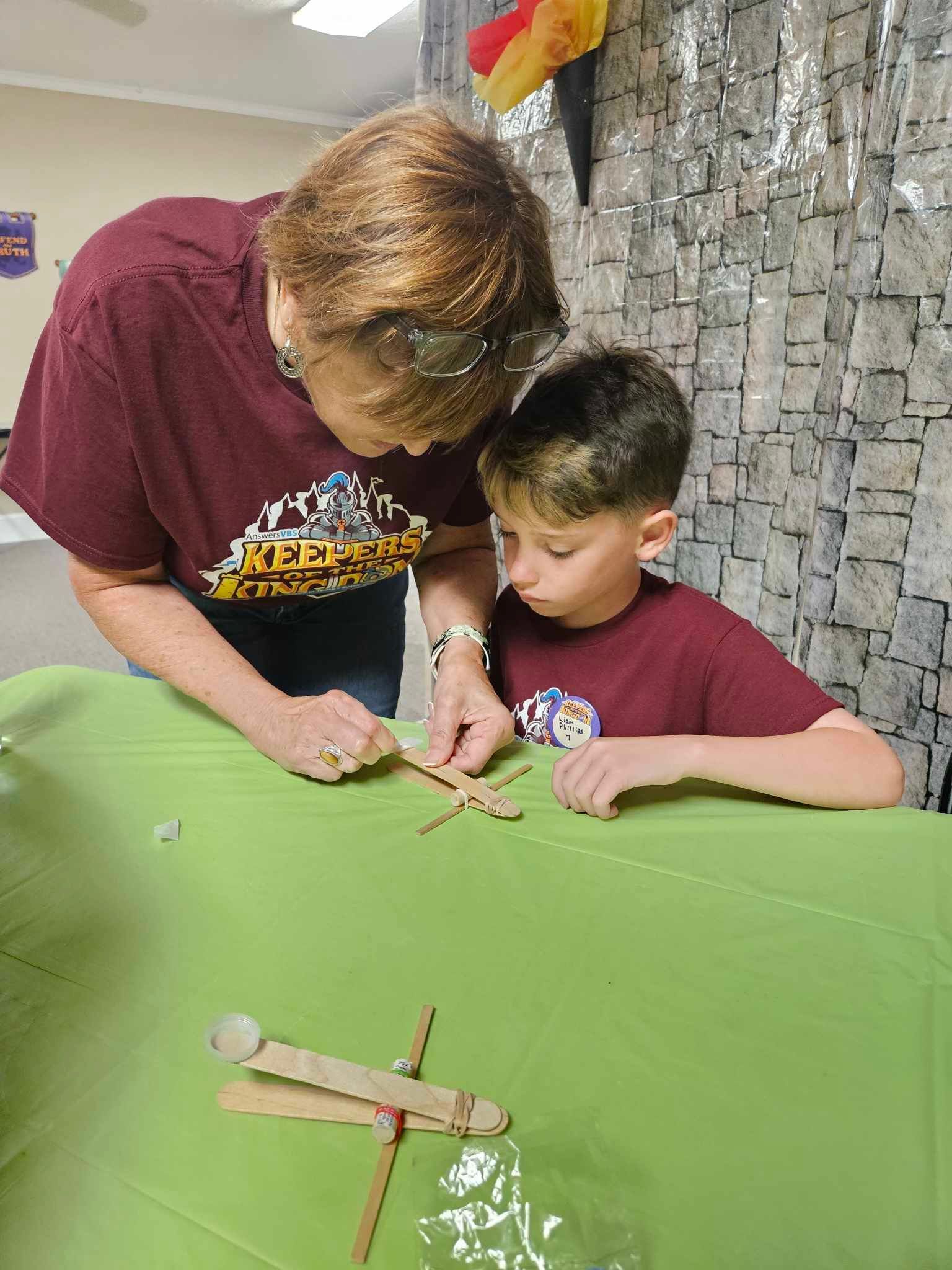 A woman and a boy are sitting at a table making a dragonfly out of popsicle sticks.