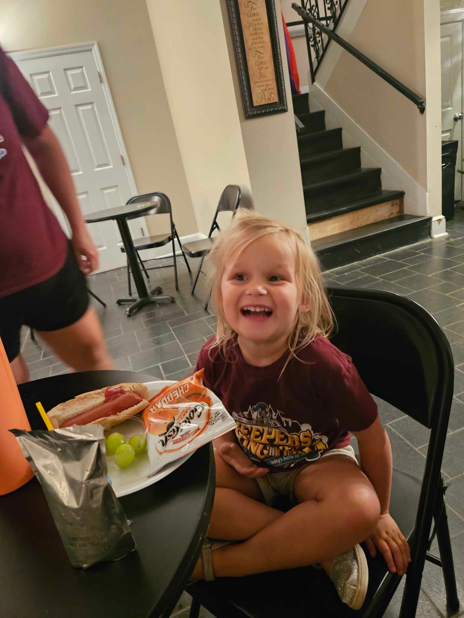 A little girl is sitting at a table with a hot dog and grapes.