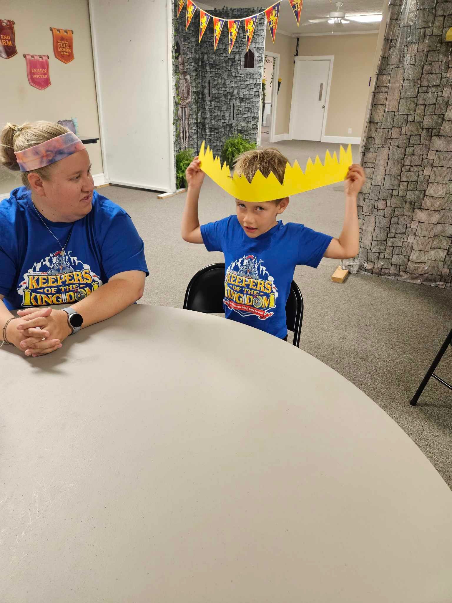 A woman is sitting at a table next to a boy wearing a crown on his head.