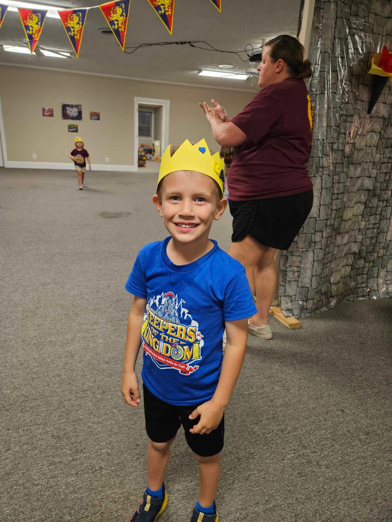 A young boy wearing a crown and a blue shirt is standing in a room.