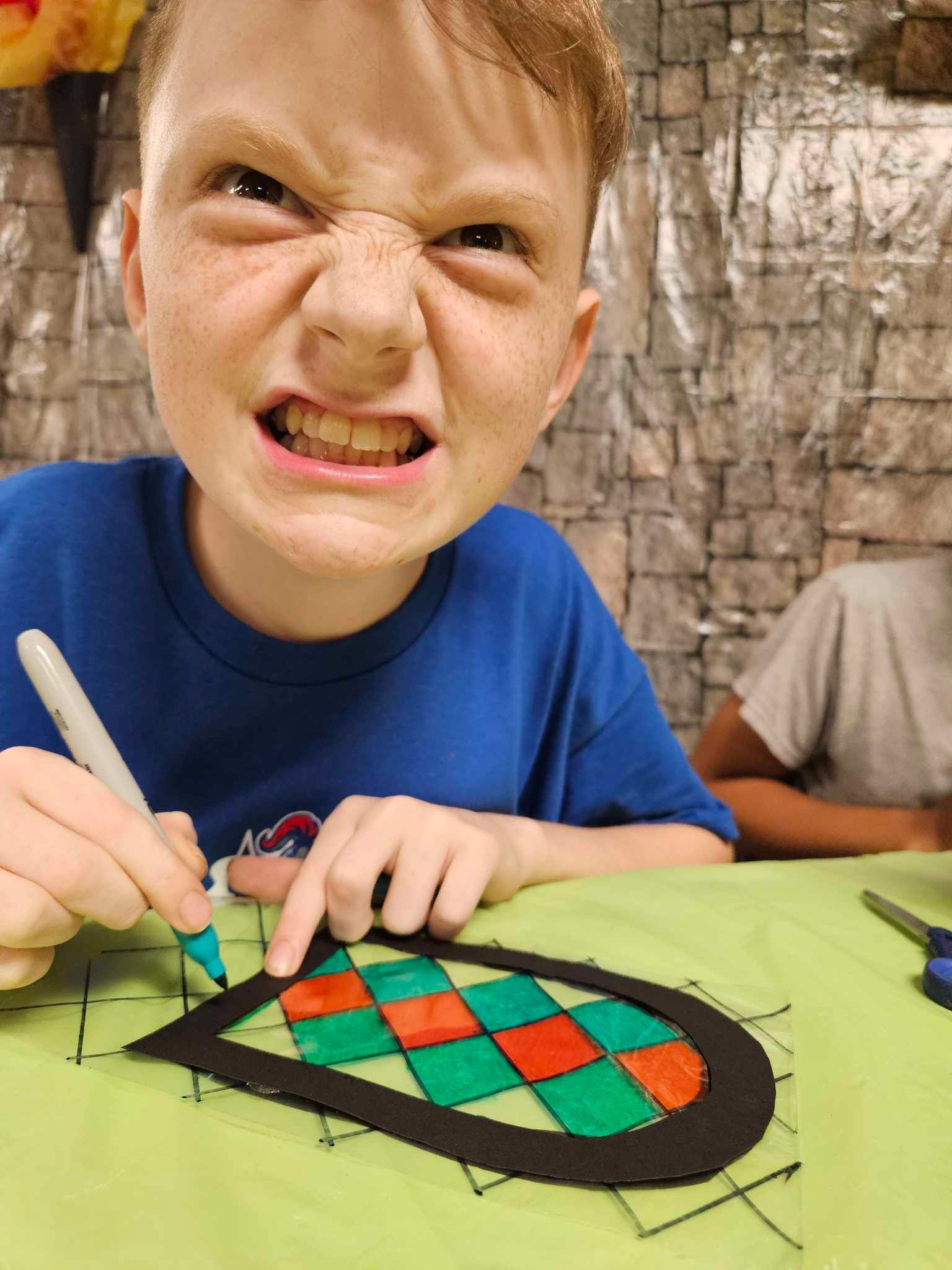 A young boy is making a funny face while drawing on a piece of paper.