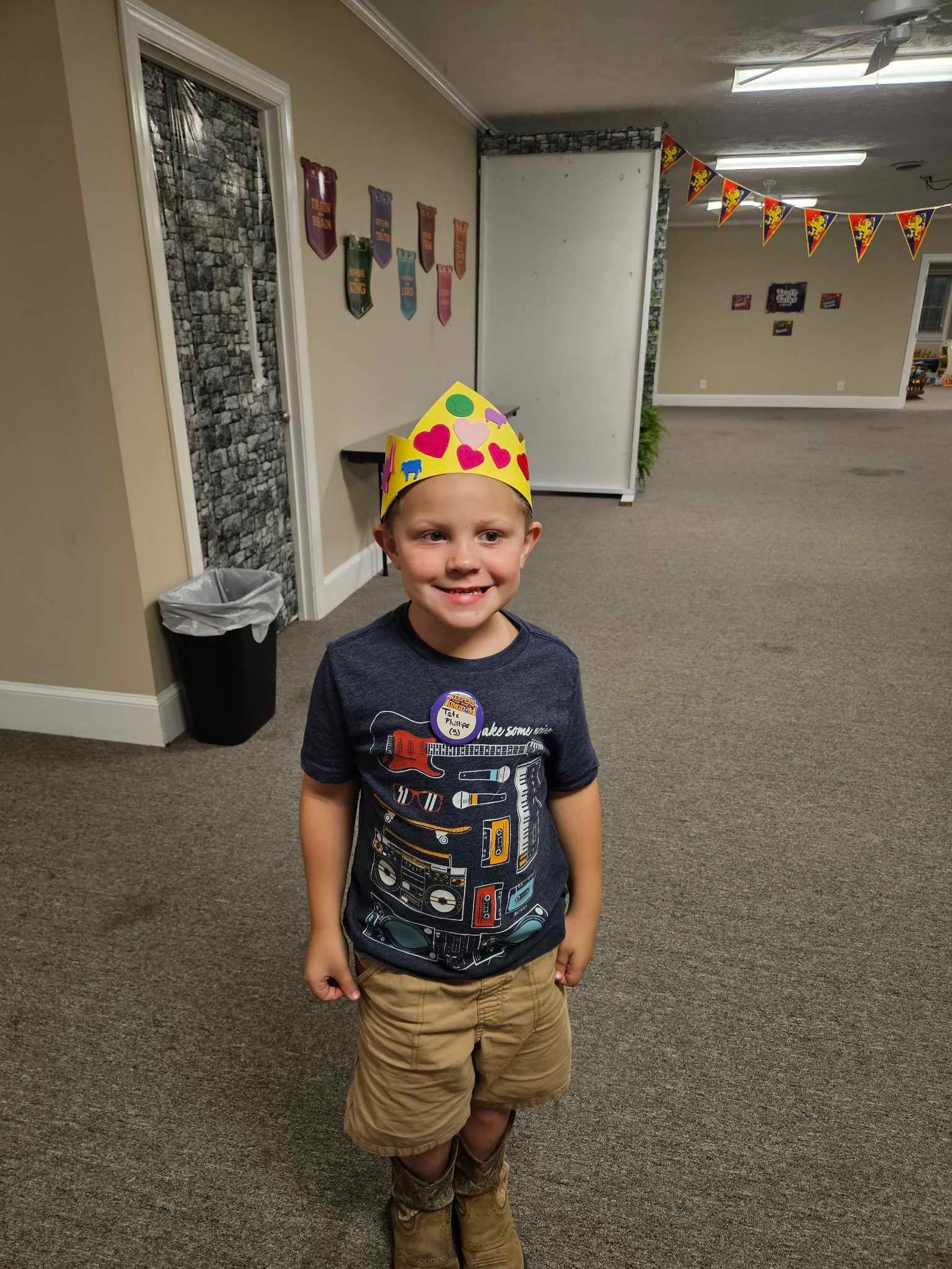 A young boy wearing a birthday hat is standing in a hallway.
