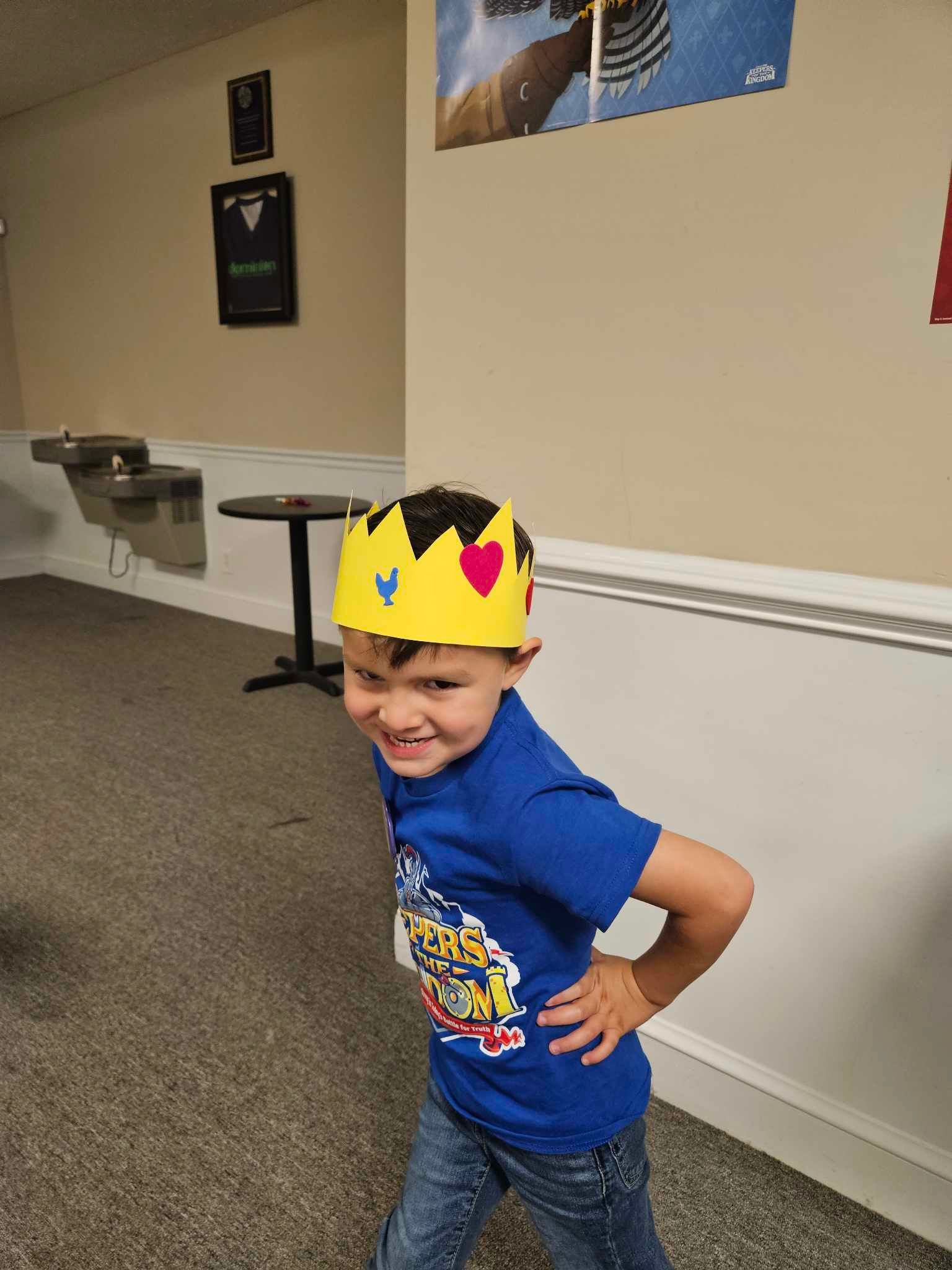 A young boy wearing a crown and a blue shirt is standing in a room.