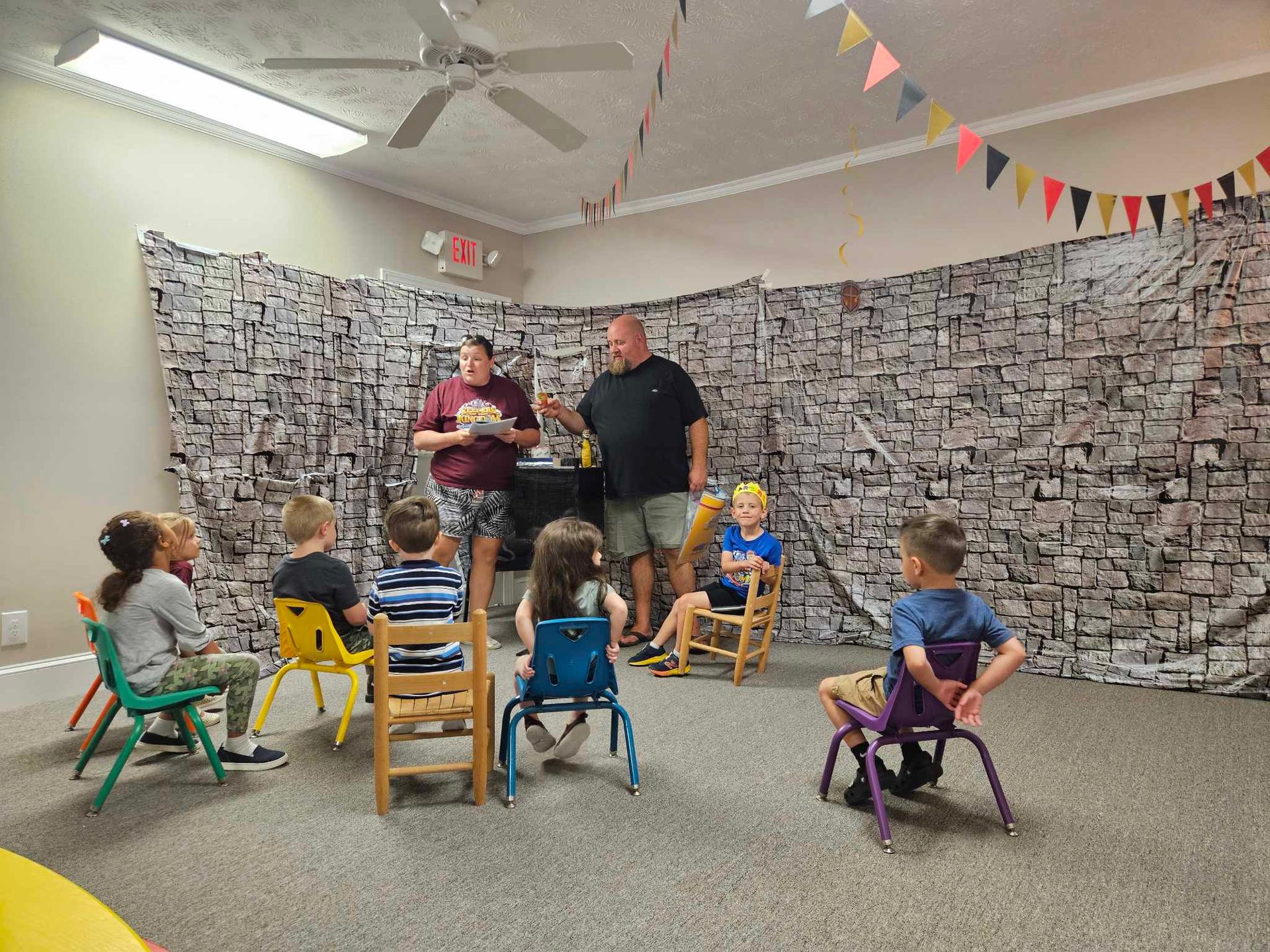 A group of children are sitting in chairs in a room with a man standing in front of them.