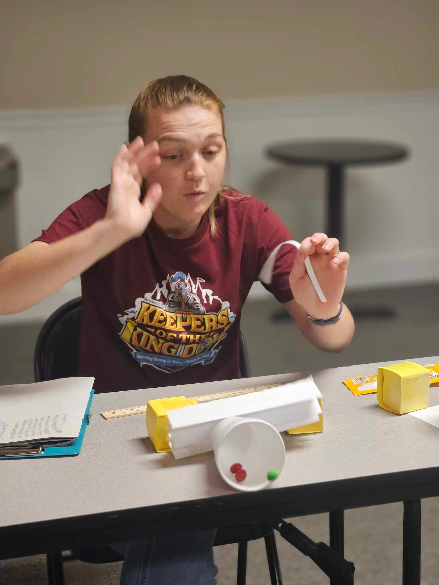 A girl in a red shirt is sitting at a table holding a piece of paper.