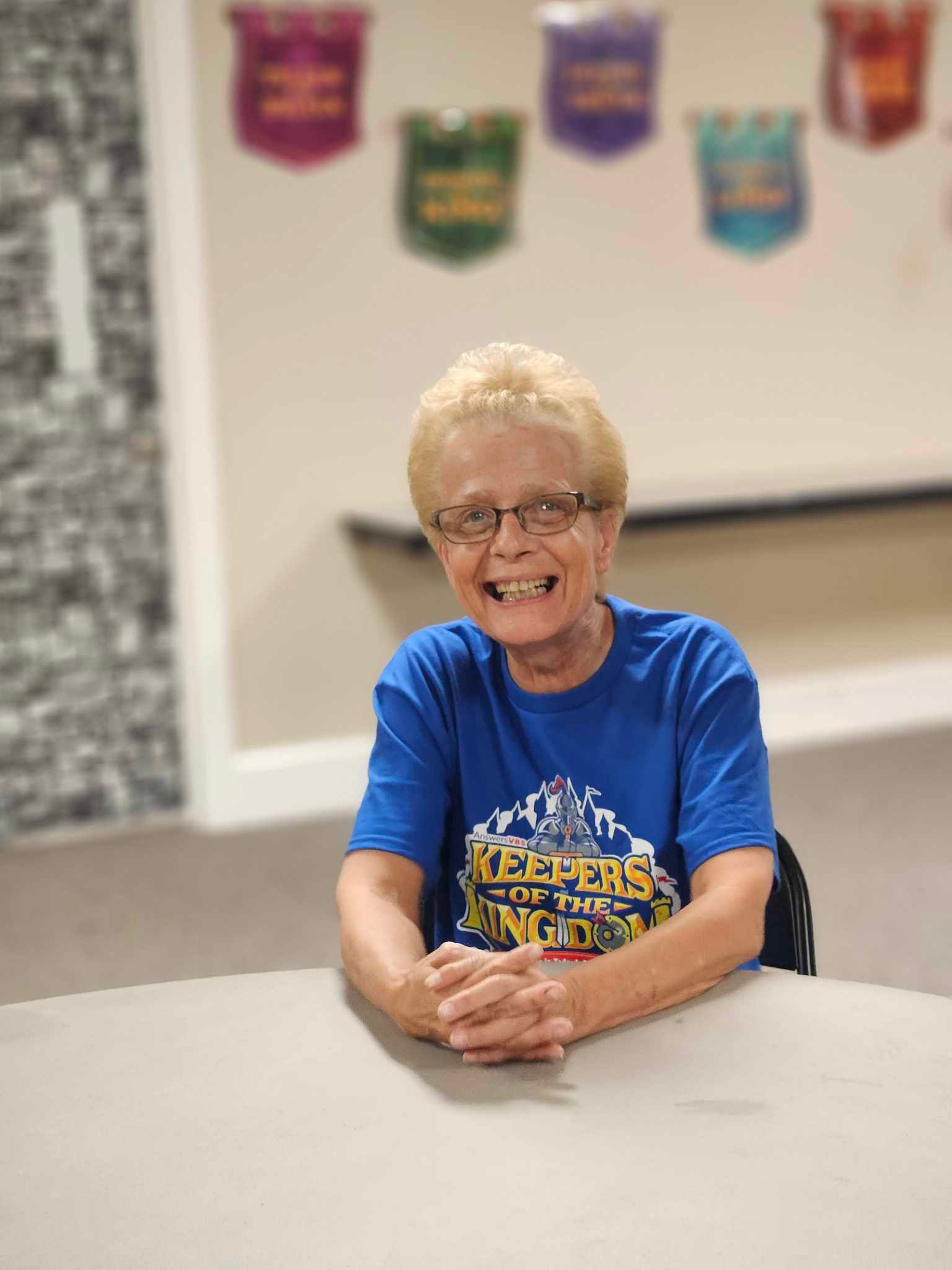 A woman in a blue shirt is sitting at a table with her hands folded and smiling.