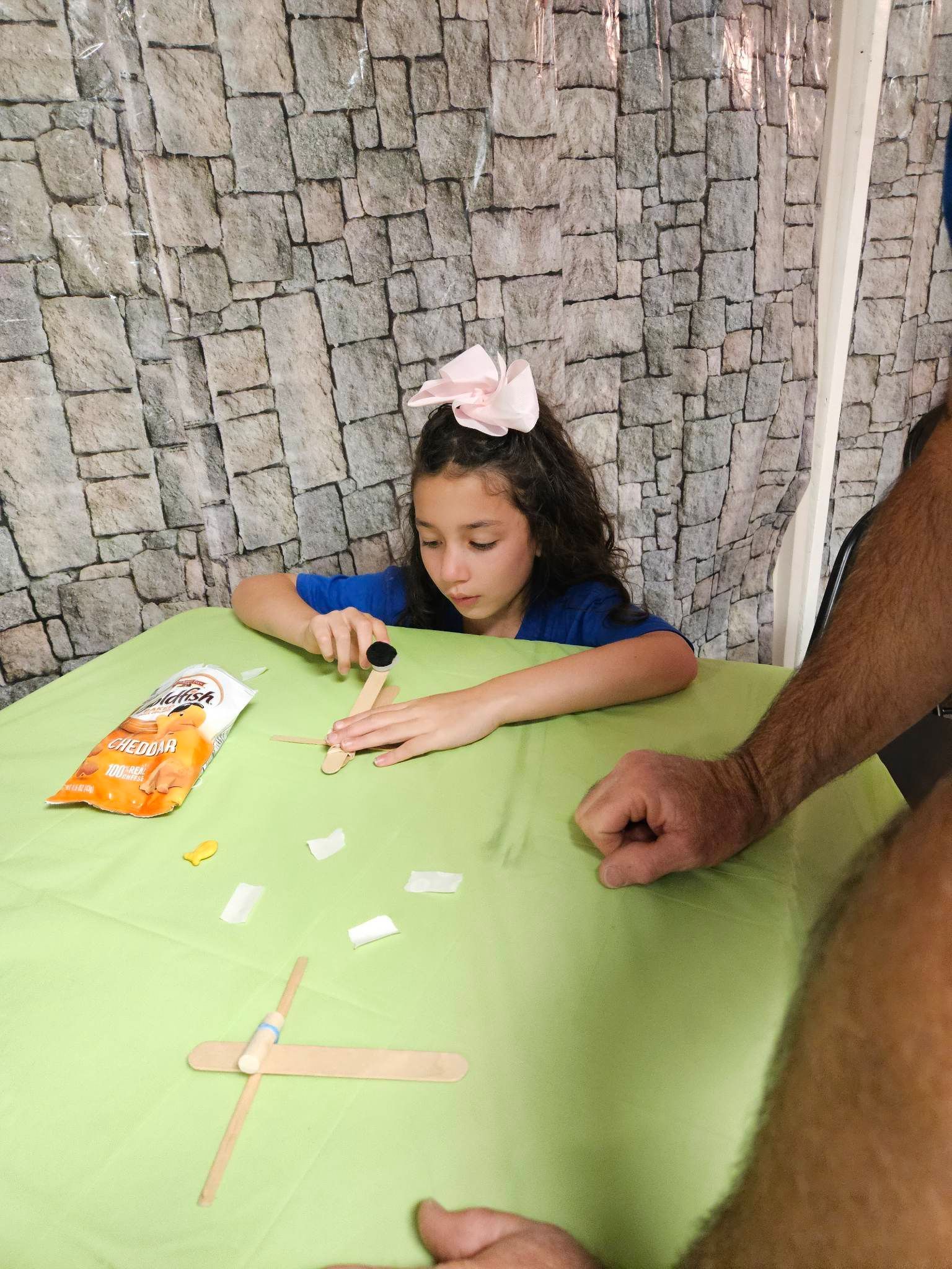 A little girl is sitting at a table making a cross out of popsicle sticks.