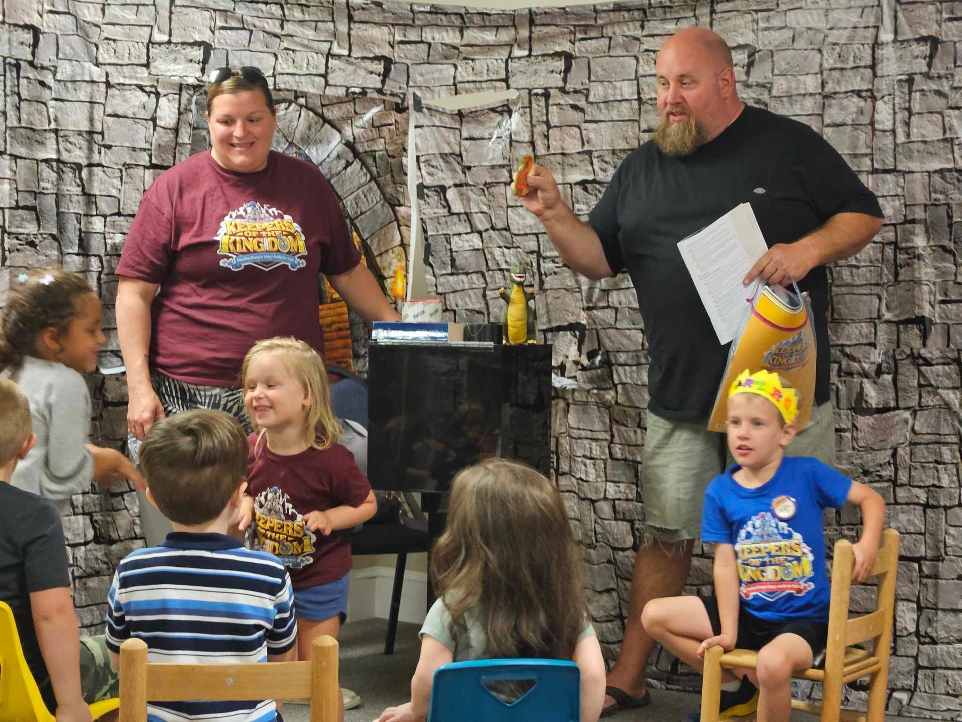 A group of children are sitting in chairs in front of a man holding a piece of paper.