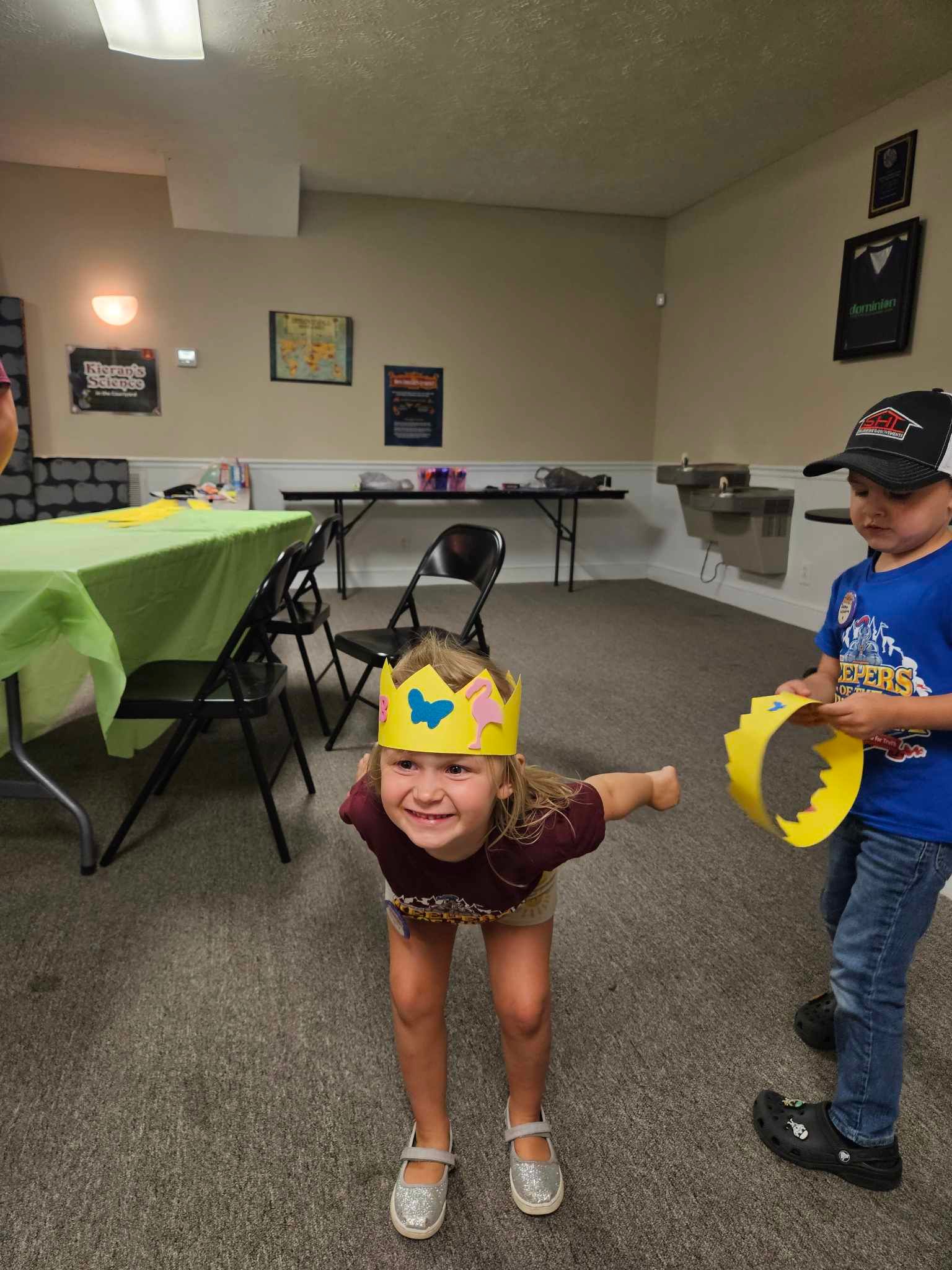 A little girl wearing a crown is standing next to a boy wearing a crown in a room.