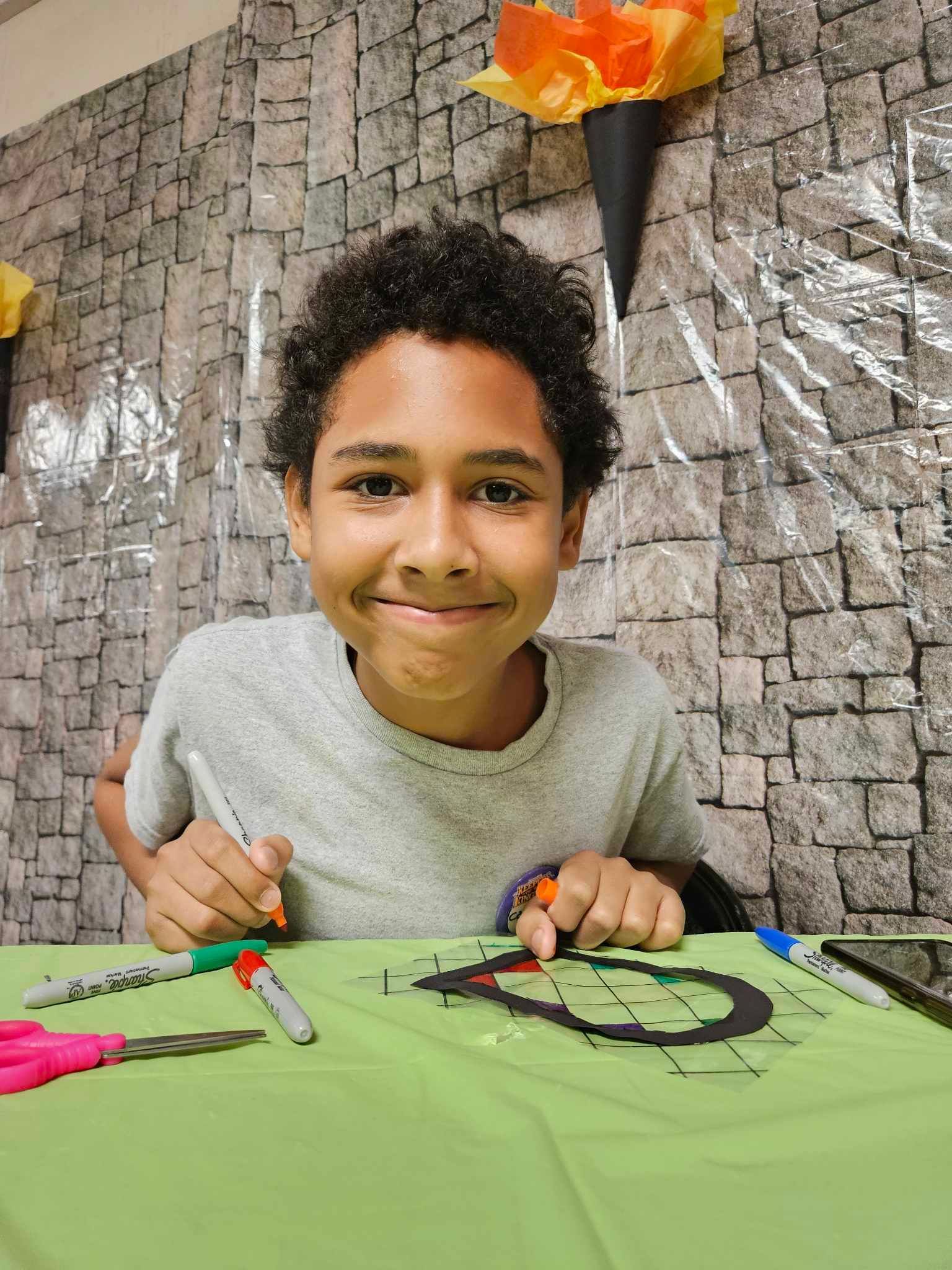 A young boy is sitting at a table with scissors and markers.