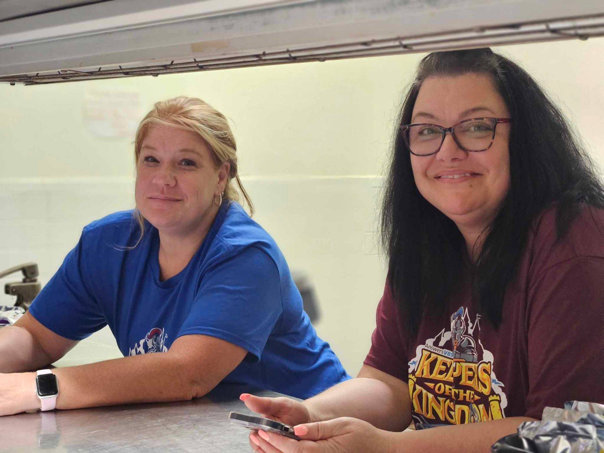 Two women are sitting at a counter with one wearing a shirt that says kings
