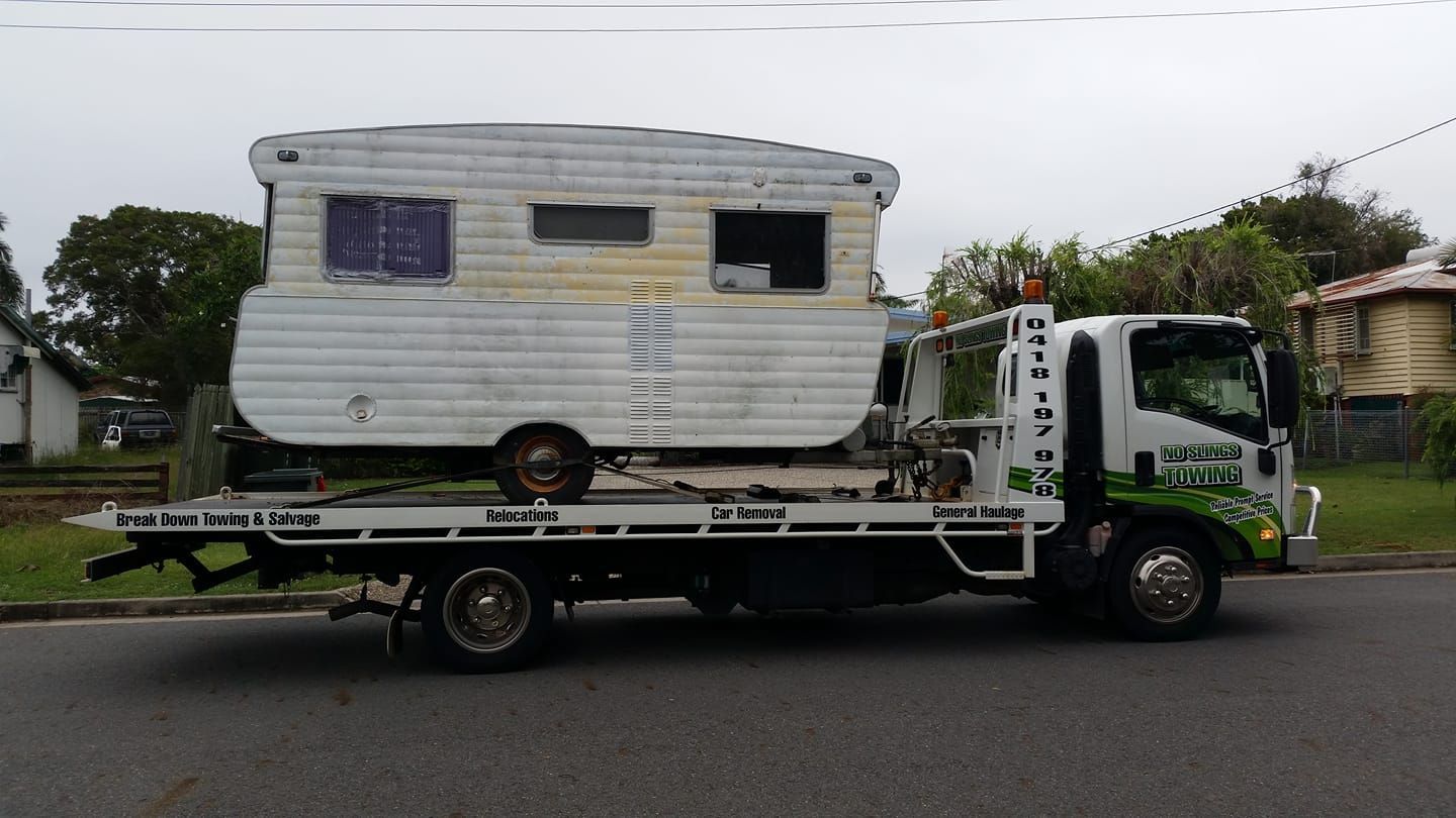 A White Tow Truck Carrying an Old, Weathered Caravan on a Street — No Slings Towing in Glendale, QLD