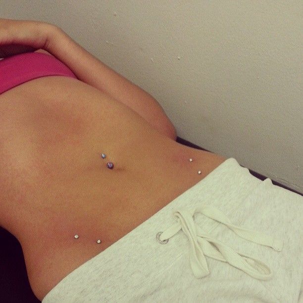 A woman in a pink bra and white shorts has multiple piercings on her stomach