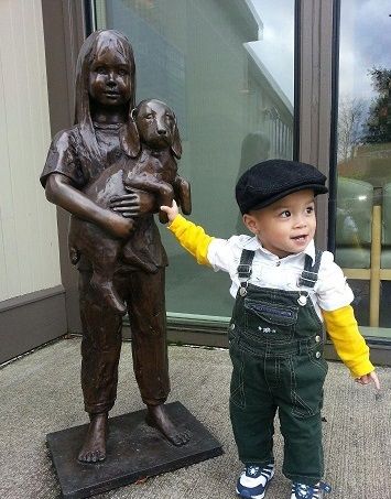 A toddler in overalls reaches towards a bronze statue of a girl holding a dog outside a building.