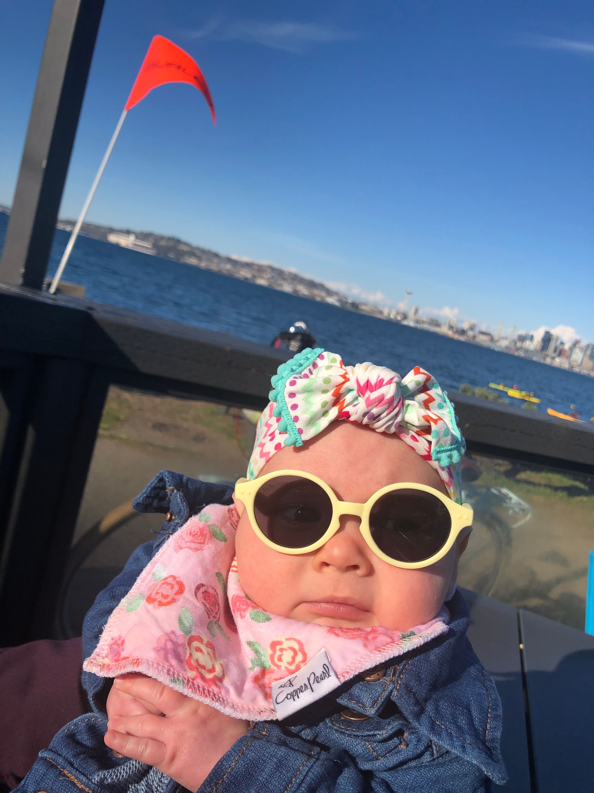 Baby wearing sunglasses and a headband, looking at the camera, with ocean and city in the background.