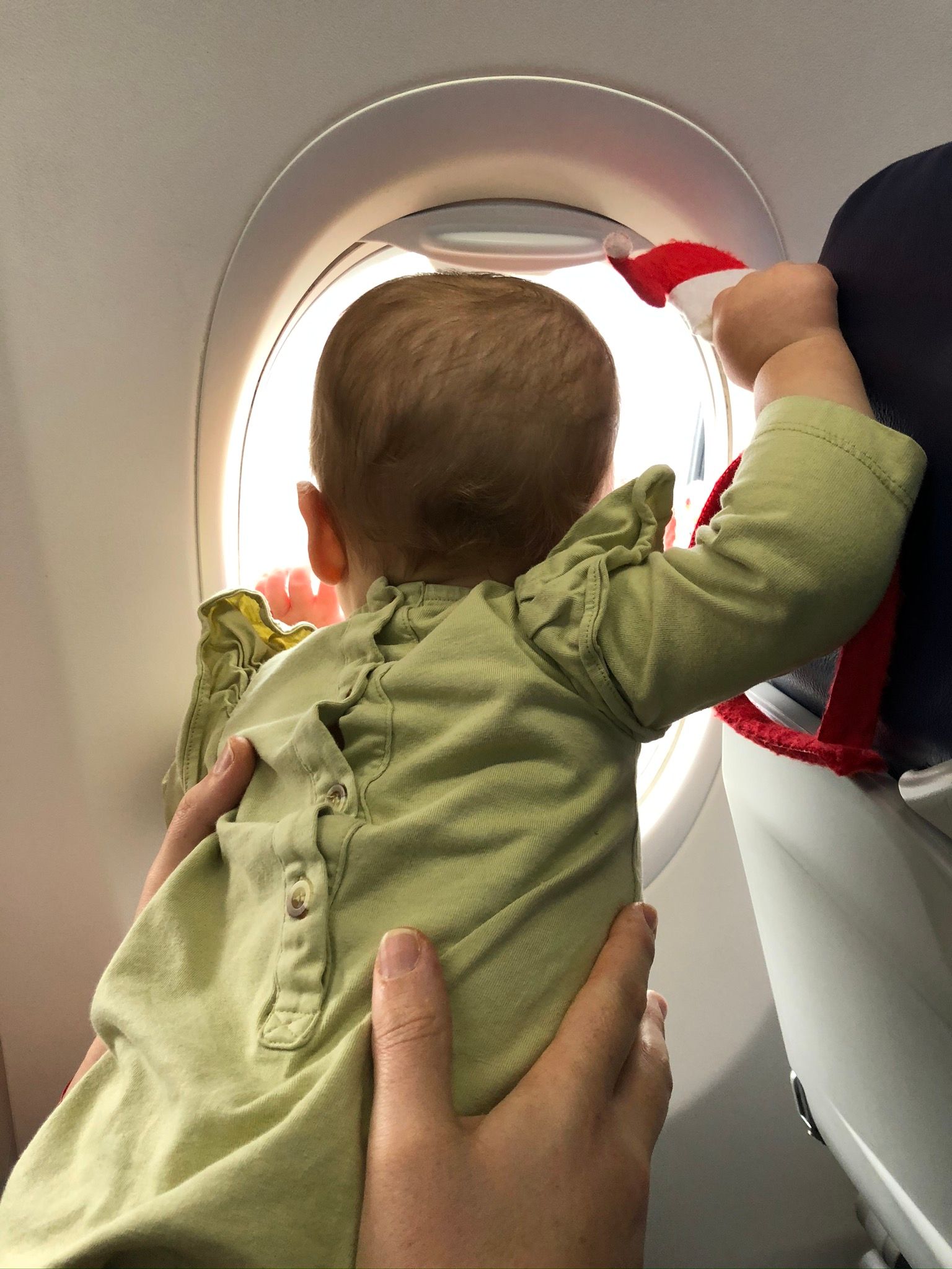 Baby looking out airplane window, held by adult. Light green outfit, bright sunlight, in-flight setting.