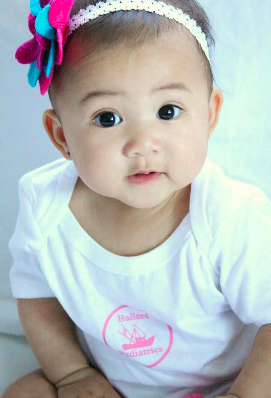 Baby wearing a white onesie and a pink/blue flower headband.