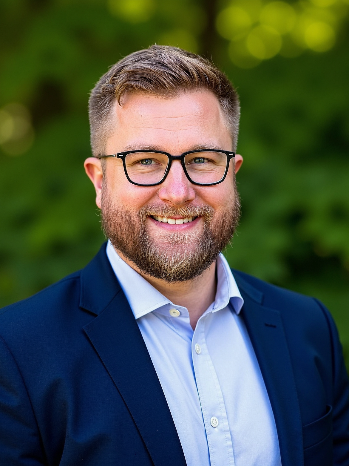 Man with beard wearing glasses, blue blazer, smiling against a blurred green background.