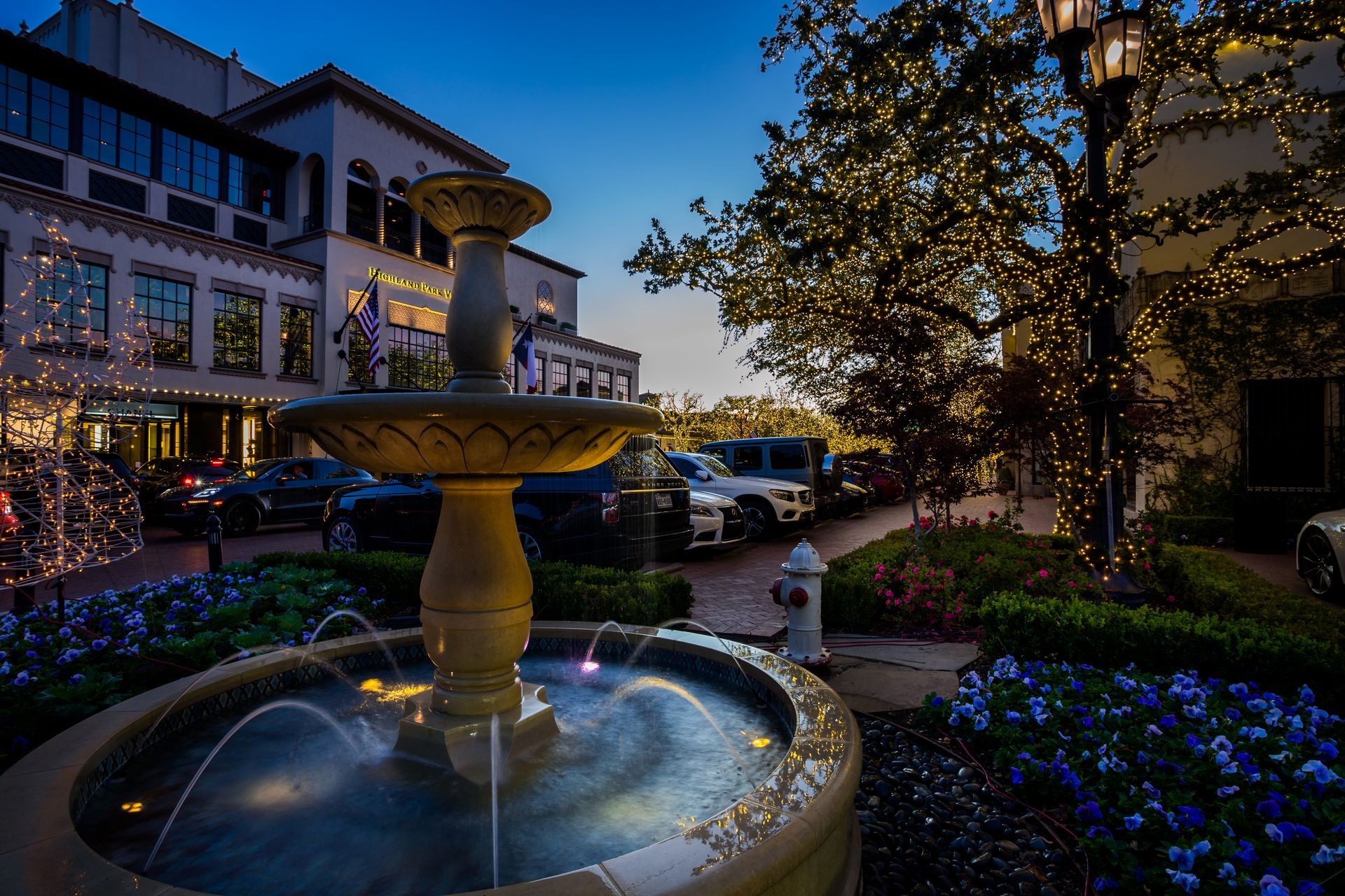 Fountain in courtyard at dusk, with illuminated buildings, trees, and parked cars.