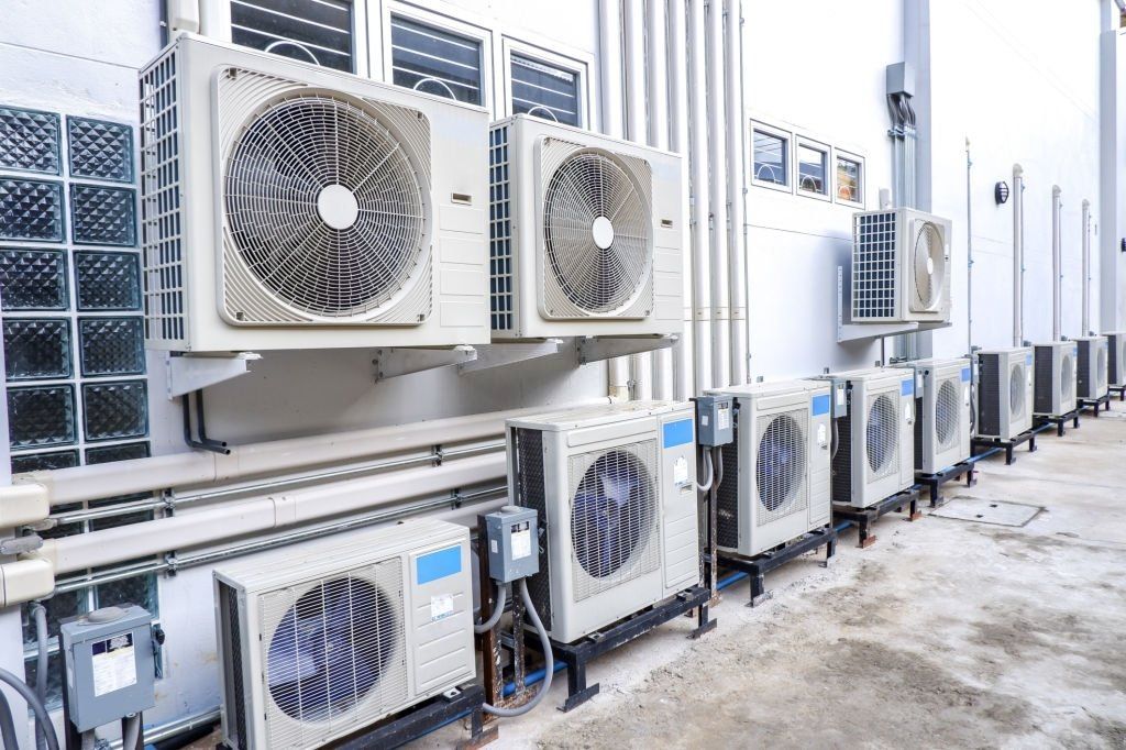 Row of air conditioning units mounted on the side of a white building. Some are on the wall, others on the ground.