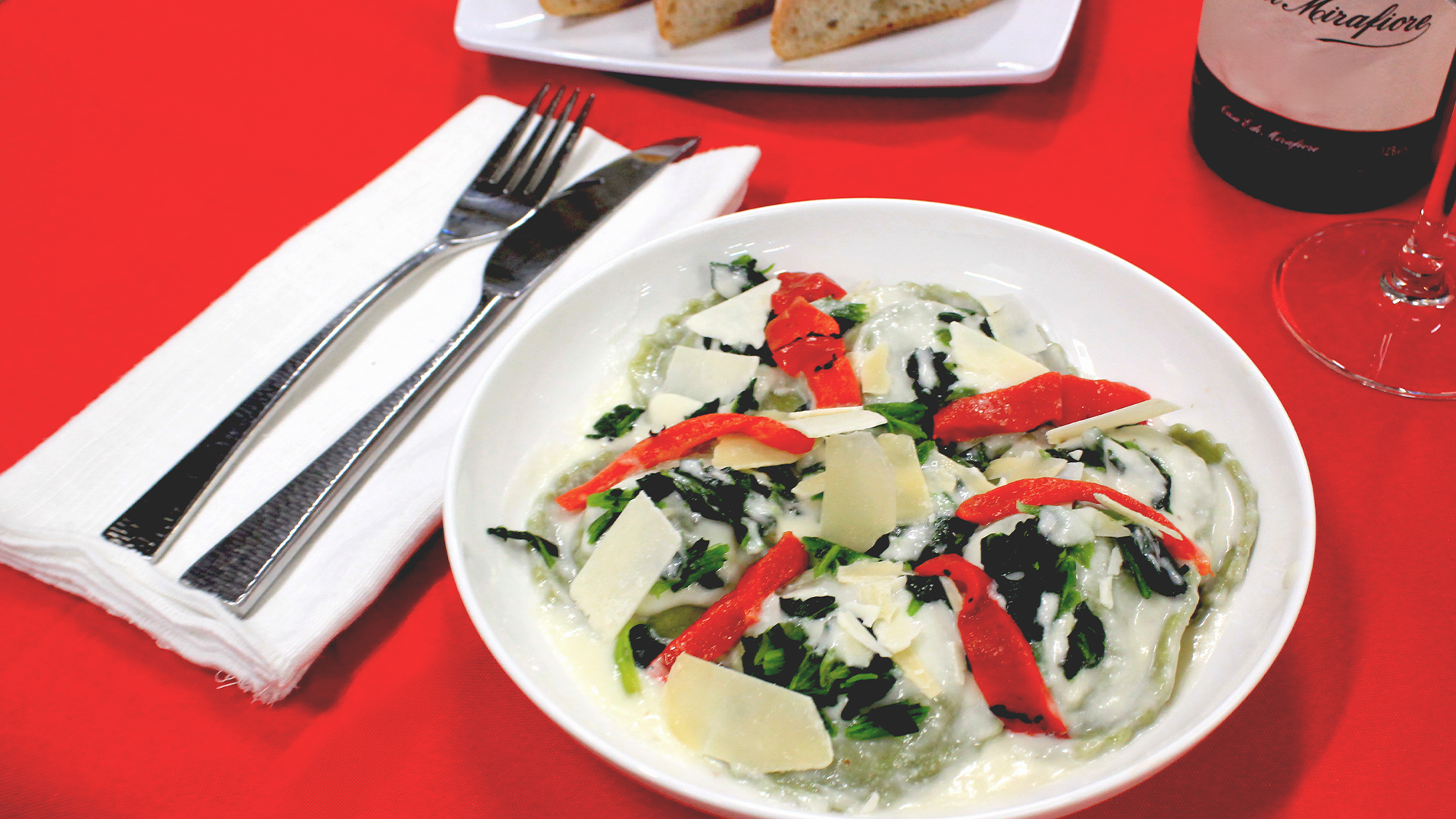 A plate of ravioli with spinach, red peppers, and parmesan on a red tablecloth with cutlery and bread.