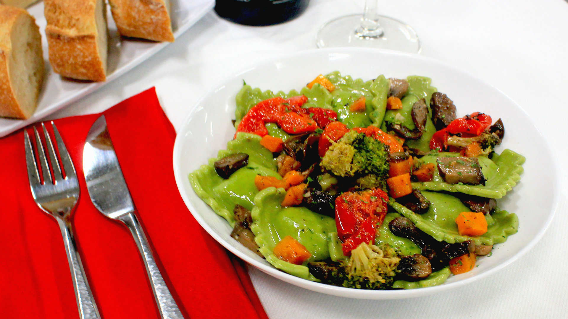 A plate of green ravioli with roasted vegetables, including broccoli and peppers, served with bread and cutlery.